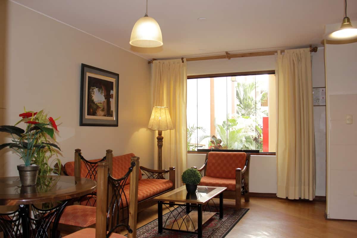 Un salon comprenant un canapé, des fauteuils, une table basse et une lampe. Il y a une fenêtre avec des rideaux, une plante et un tableau encadré accroché au mur.