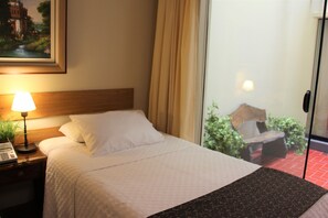 Une chambre d’hôtel avec un lit, une table de chevet avec une lampe et un téléphone, un tableau au mur, et une vue sur un balcon agrémenté de plantes et d’un banc.