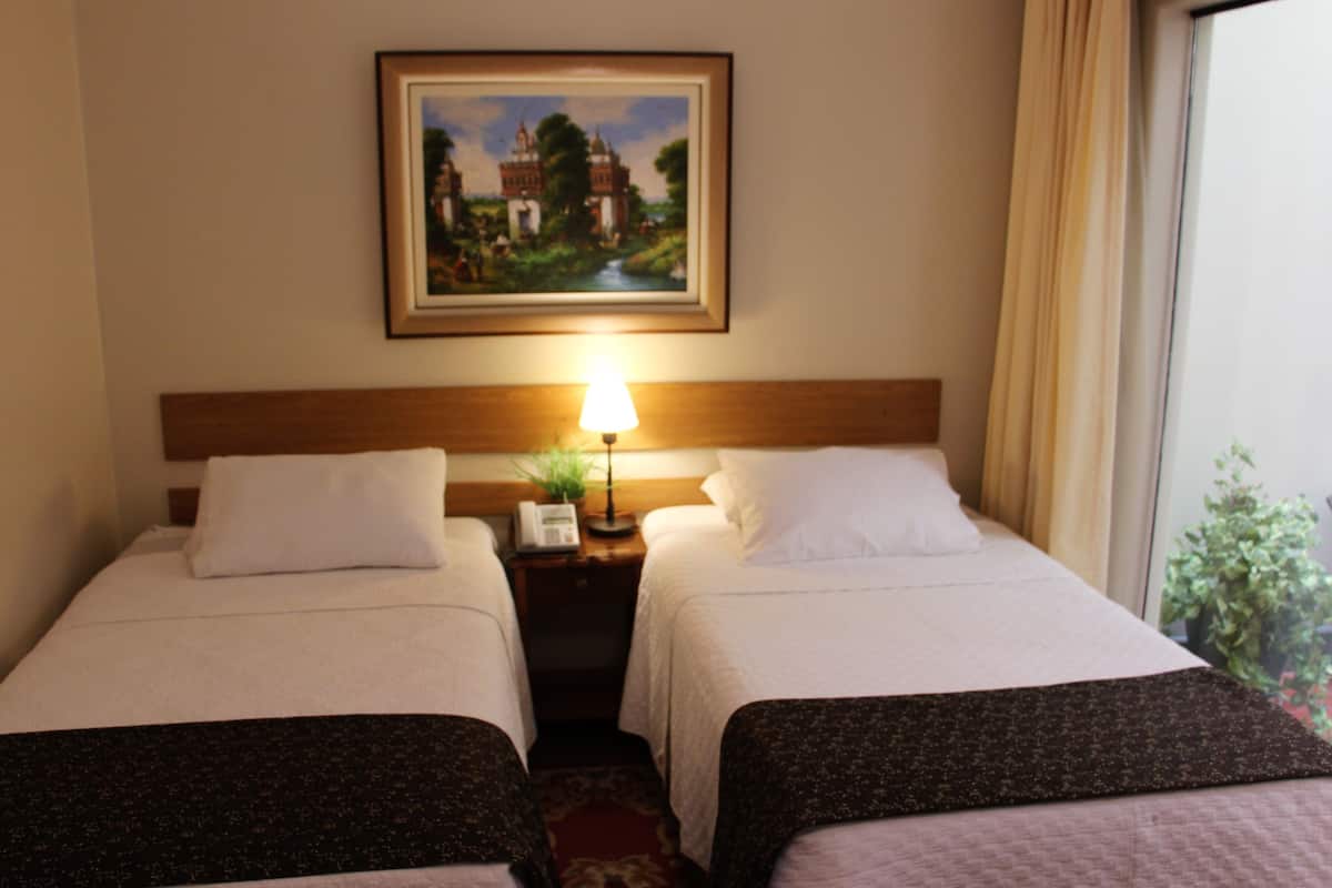 Une chambre d’hôtel avec deux lits, une table de chevet avec une lampe, un tableau au mur et une fenêtre avec des rideaux.