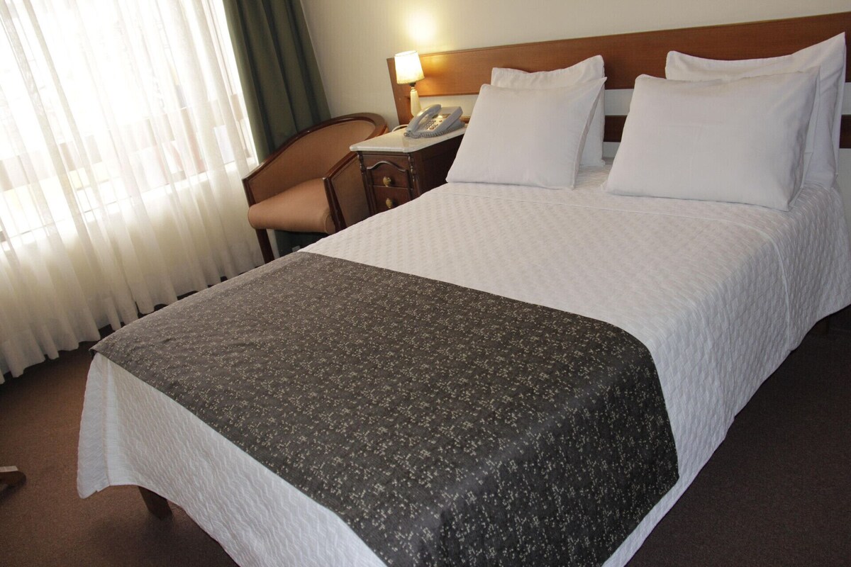 A hotel room with a bed, a chair, a nightstand with a lamp and phone, and a window with curtains.