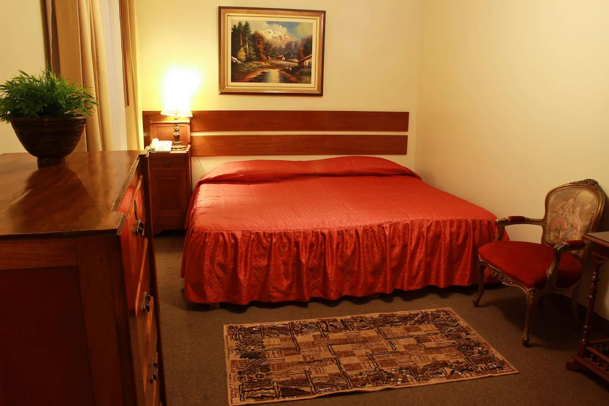 Une chambre d’hôtel avec une tête de lit en bois, une couvre-lit rouge, une table de chevet en bois avec une lampe, une chaise, un tapis et un tableau au mur.