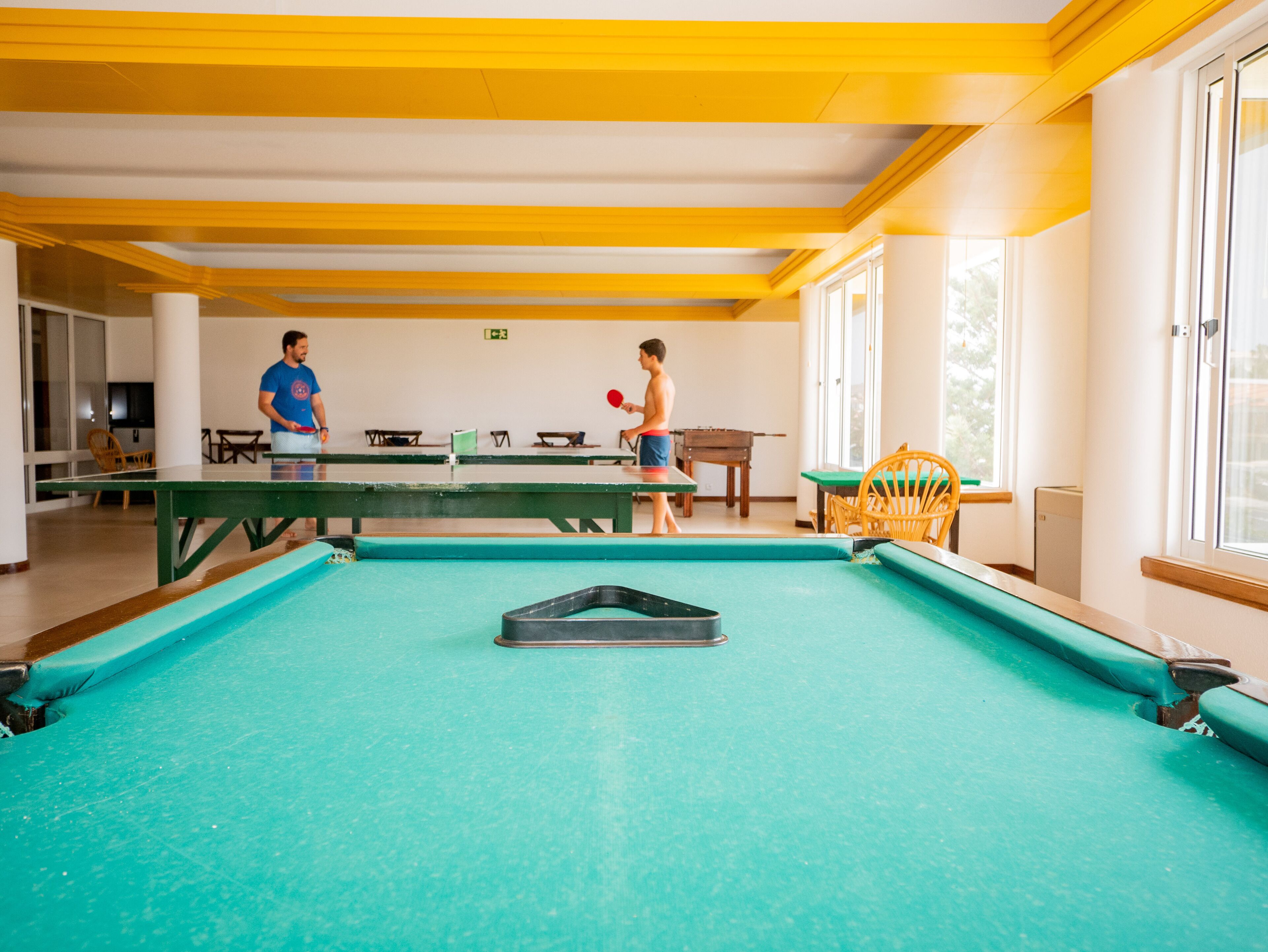 Una mesa de billar en una habitación con vigas amarillas y ventanas amplias.