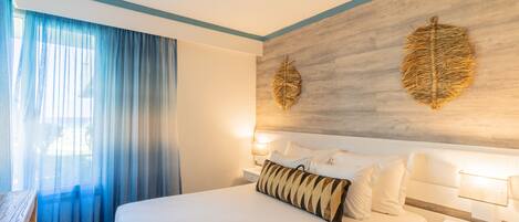A hotel room with a large bed, a wooden headboard, a nightstand with a lamp, and a window with sheer curtains.