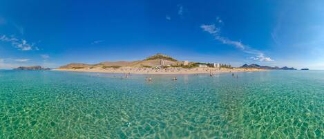 A beach with clear turquoise water, a sandy shore, and a hill with buildings.