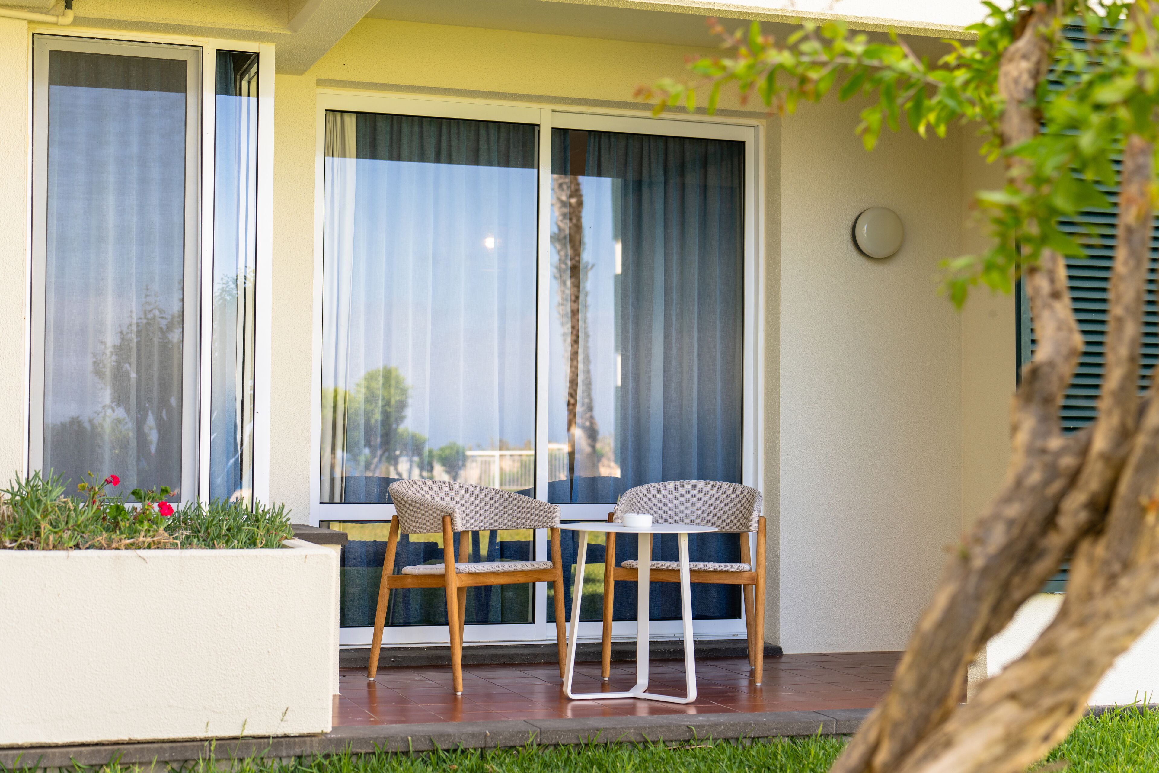Eine Terrasse mit zwei Korbstühlen und einem kleinen Tisch, großen Fenstern mit Vorhängen sowie einem Blumentopf mit roten Blüten.
