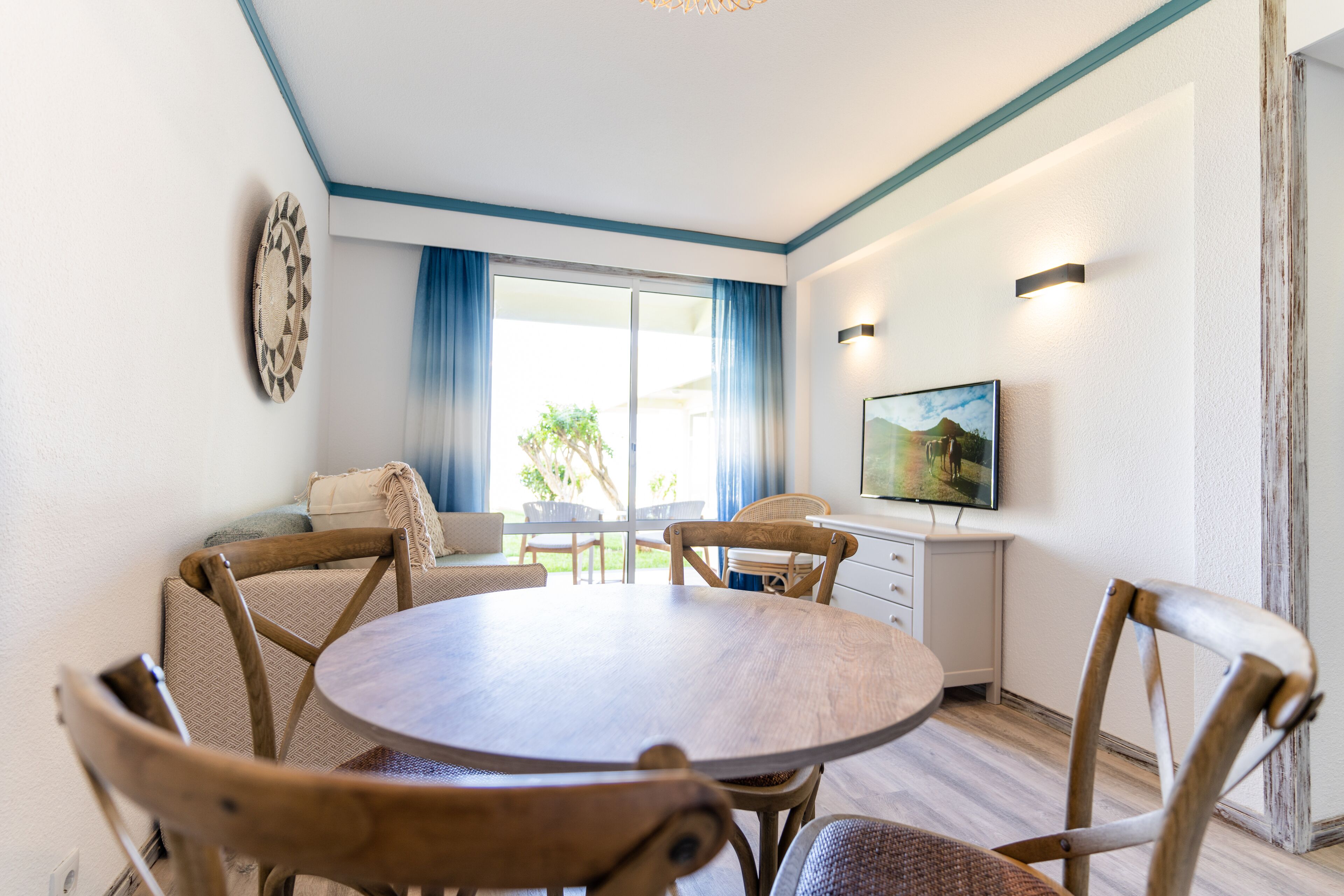A dining area with a round table and wooden chairs, a television on the wall, and a view of the outdoors through a large window with blue curtains.