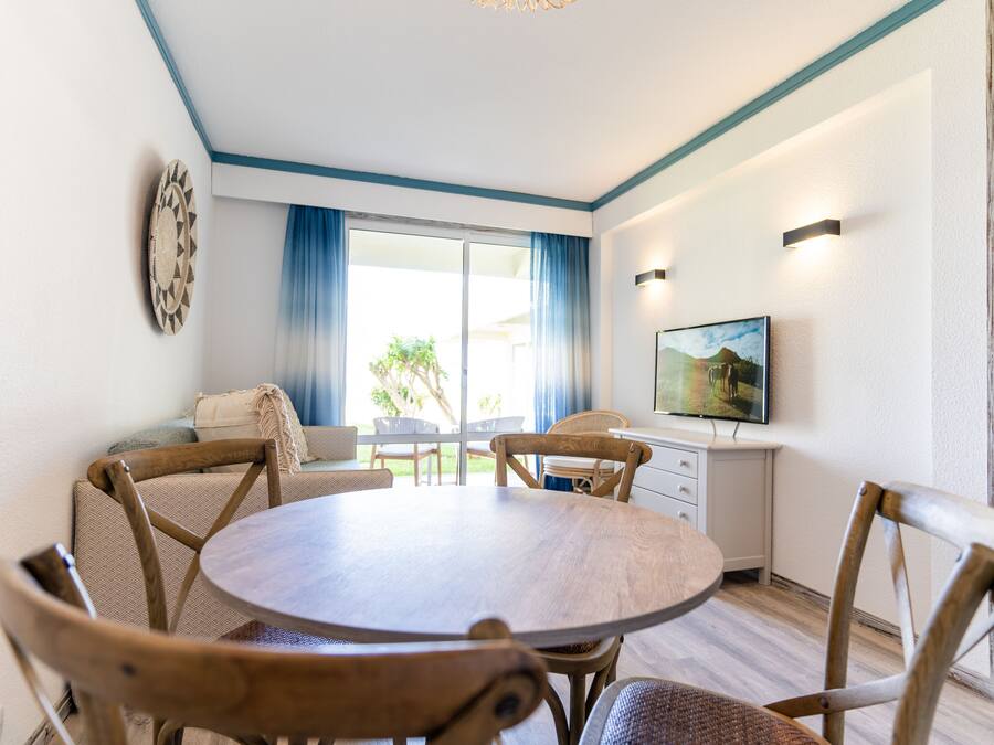 A dining area with a round table and wooden chairs, a television on the wall, and a view of the outdoors through a large window with blue curtains.