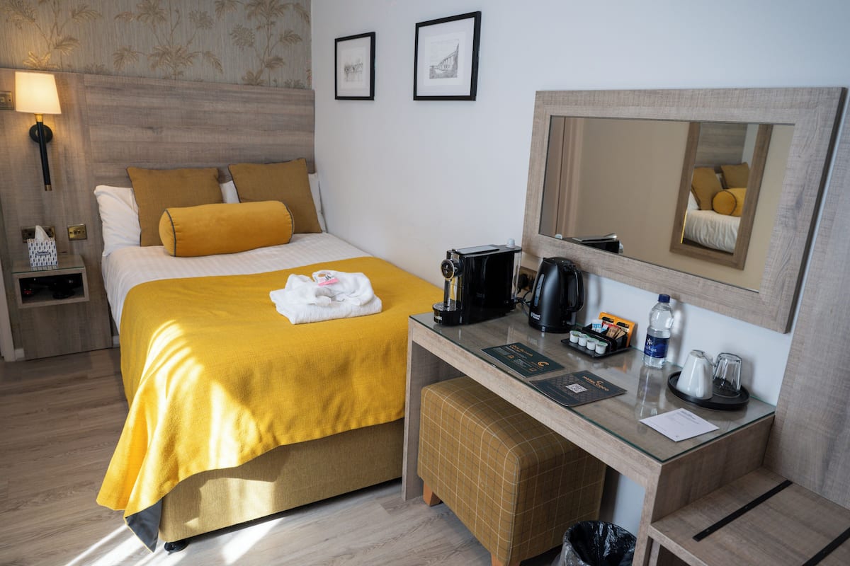 Een hotelkamer met een bed, nachtkastje, spiegel en een klein bureau met een koffiezetapparaat.