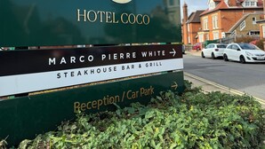 Une enseigne d’hôtel avec un logo, le nom de l’établissement et des indications pour se rendre à un bar-restaurant spécialisé dans les steaks et les grillades.