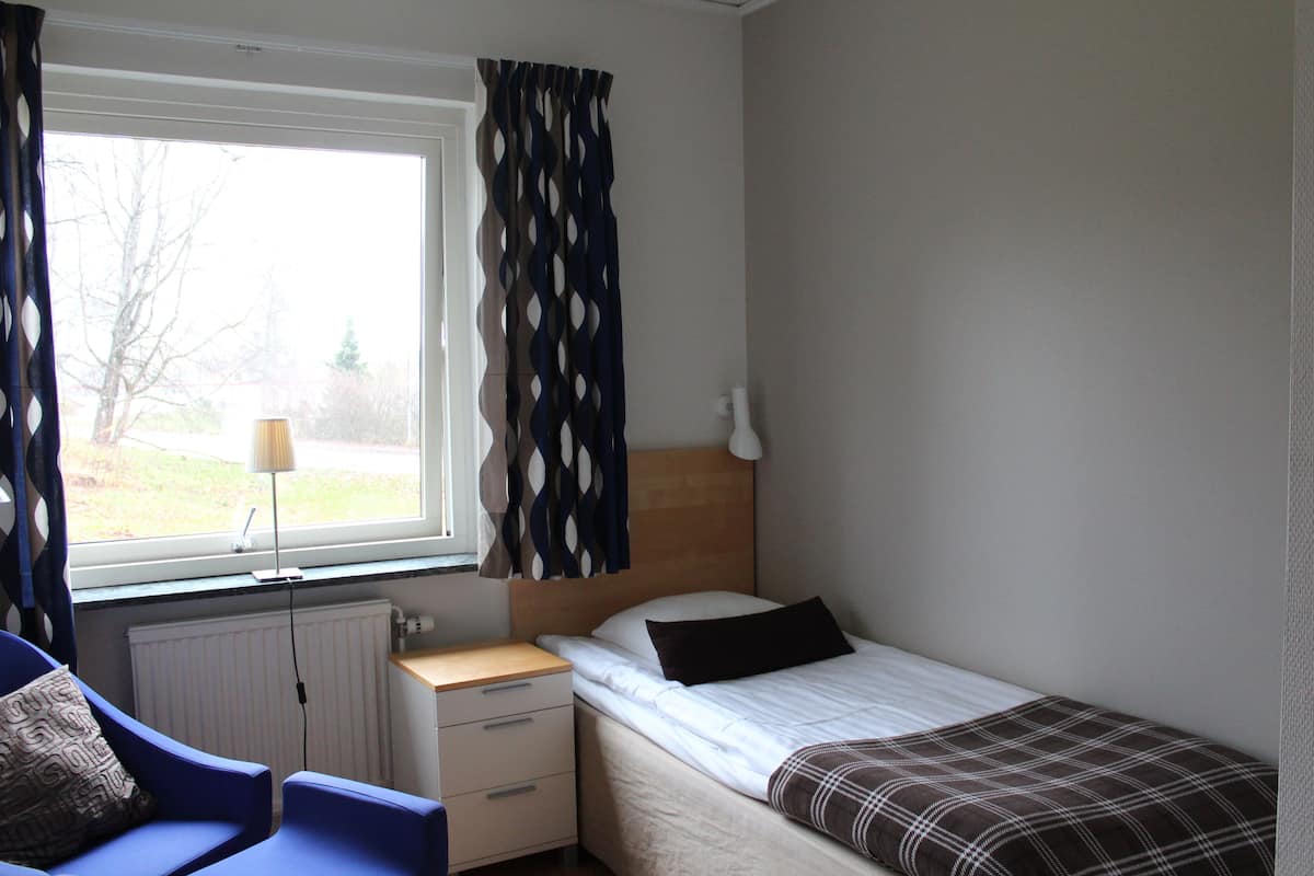 A bedroom with a bed, a nightstand, a chair, a window with curtains, and a radiator.