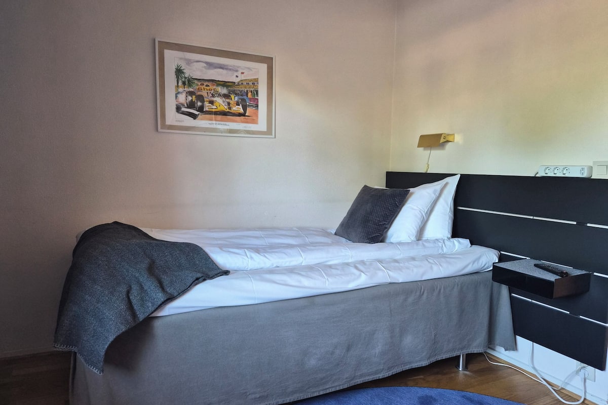 A bed with grey bedding, a black headboard, a framed picture on the wall, and a bedside table with a lamp.