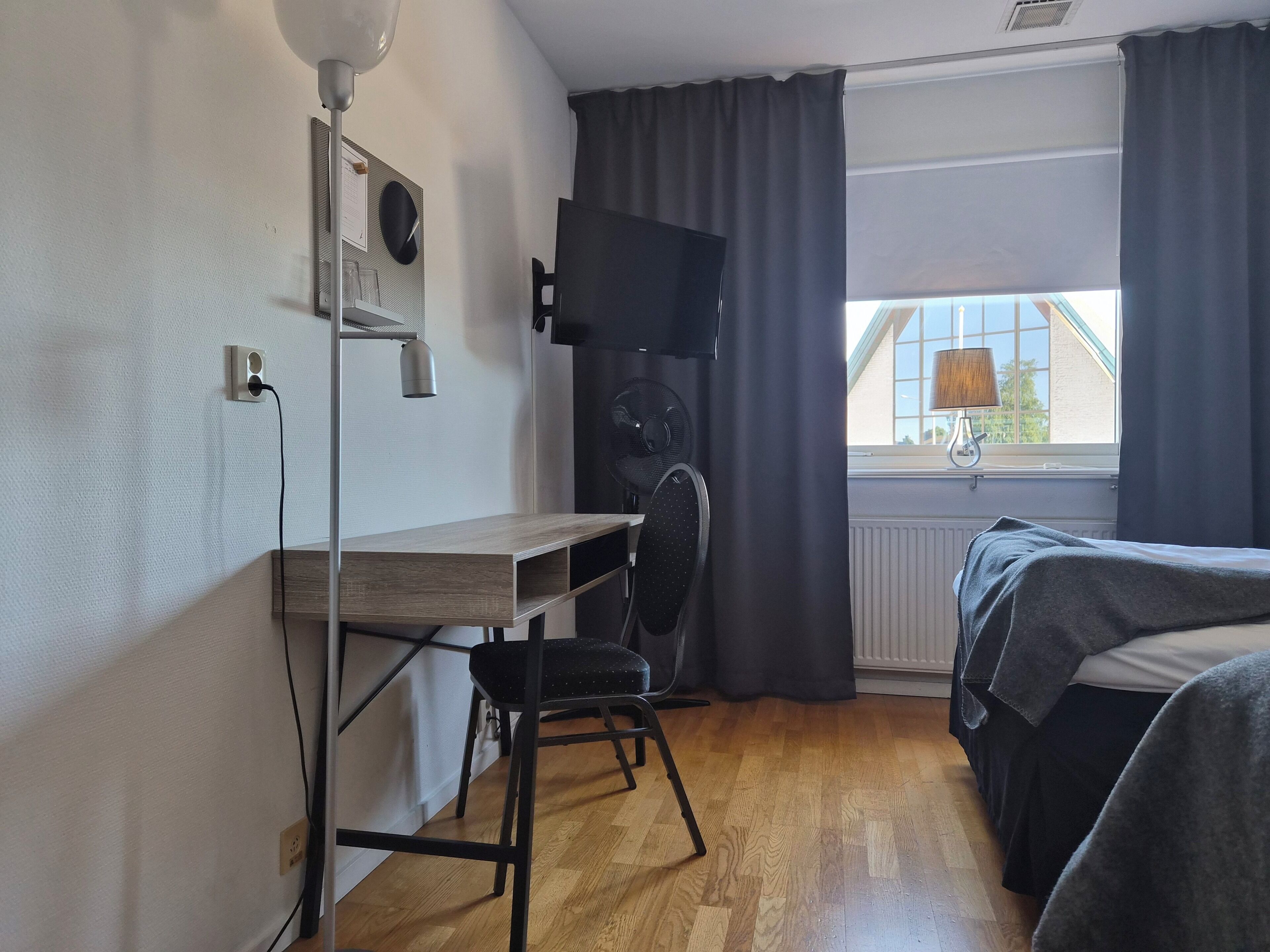 A room with a desk, chair, TV, bed, and a window with curtains.