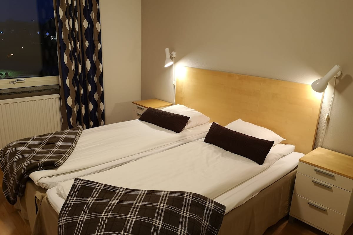 A double bed with a headboard, two bedside tables with lamps, a window with curtains, and a radiator.