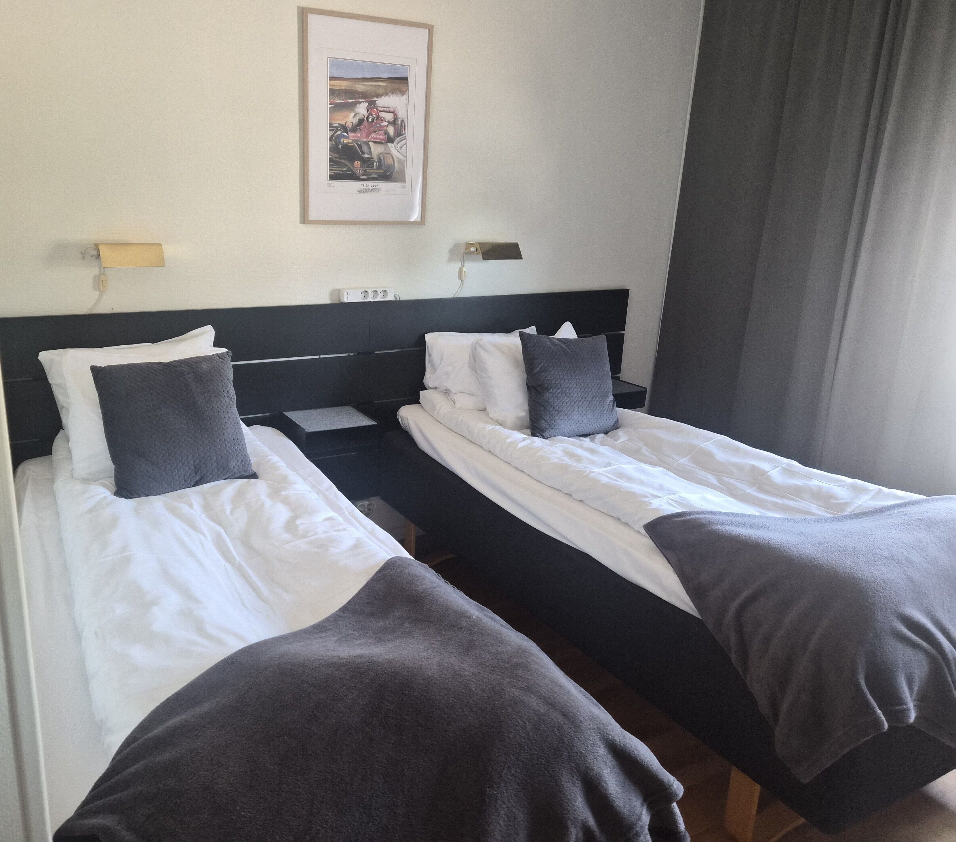 A hotel room with two single beds, a headboard, bedside tables, a wall-mounted light, and a framed picture.
