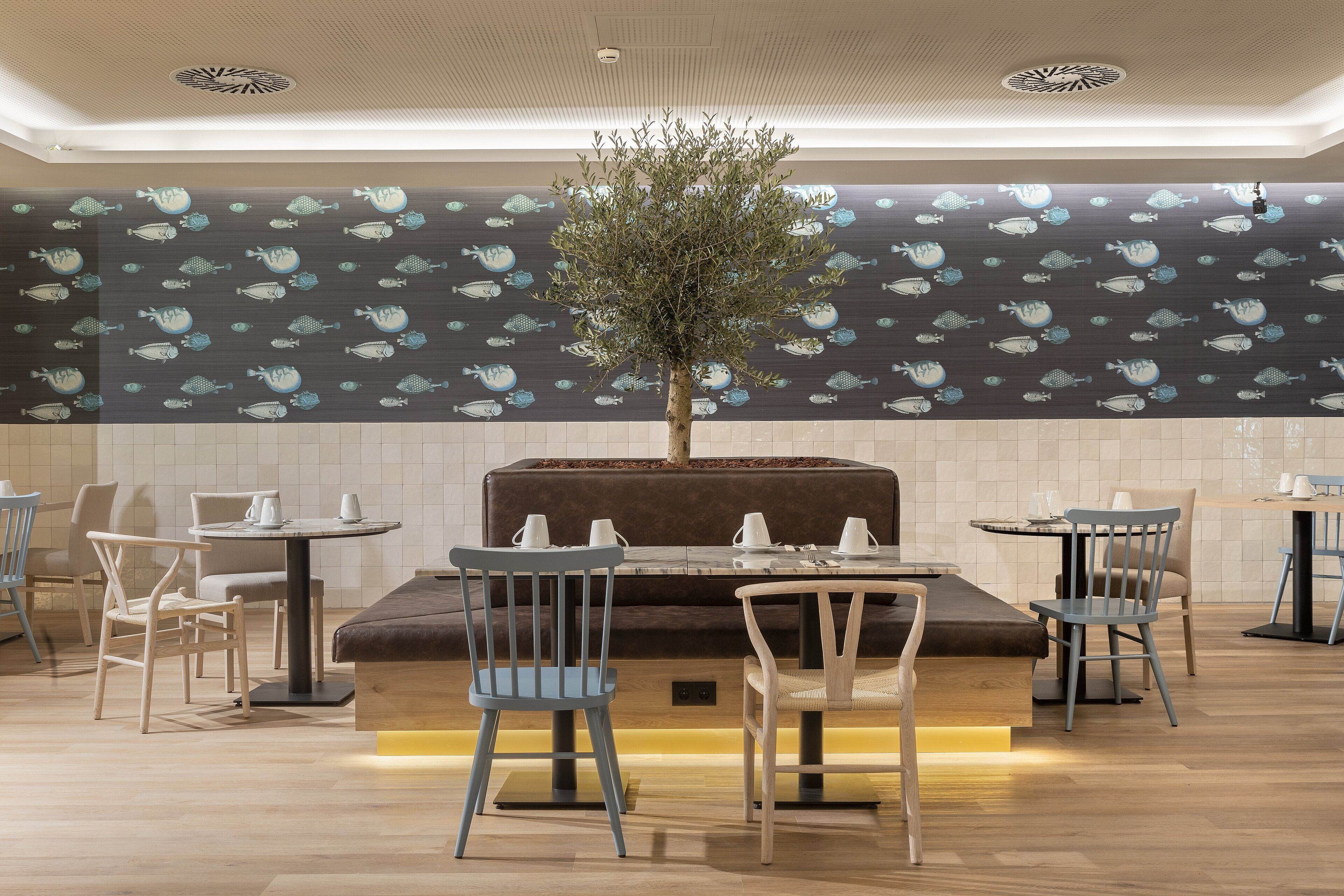 A restaurant with a central seating area featuring a brown cushioned bench, surrounded by wooden chairs. The walls are decorated with a pattern of fish.