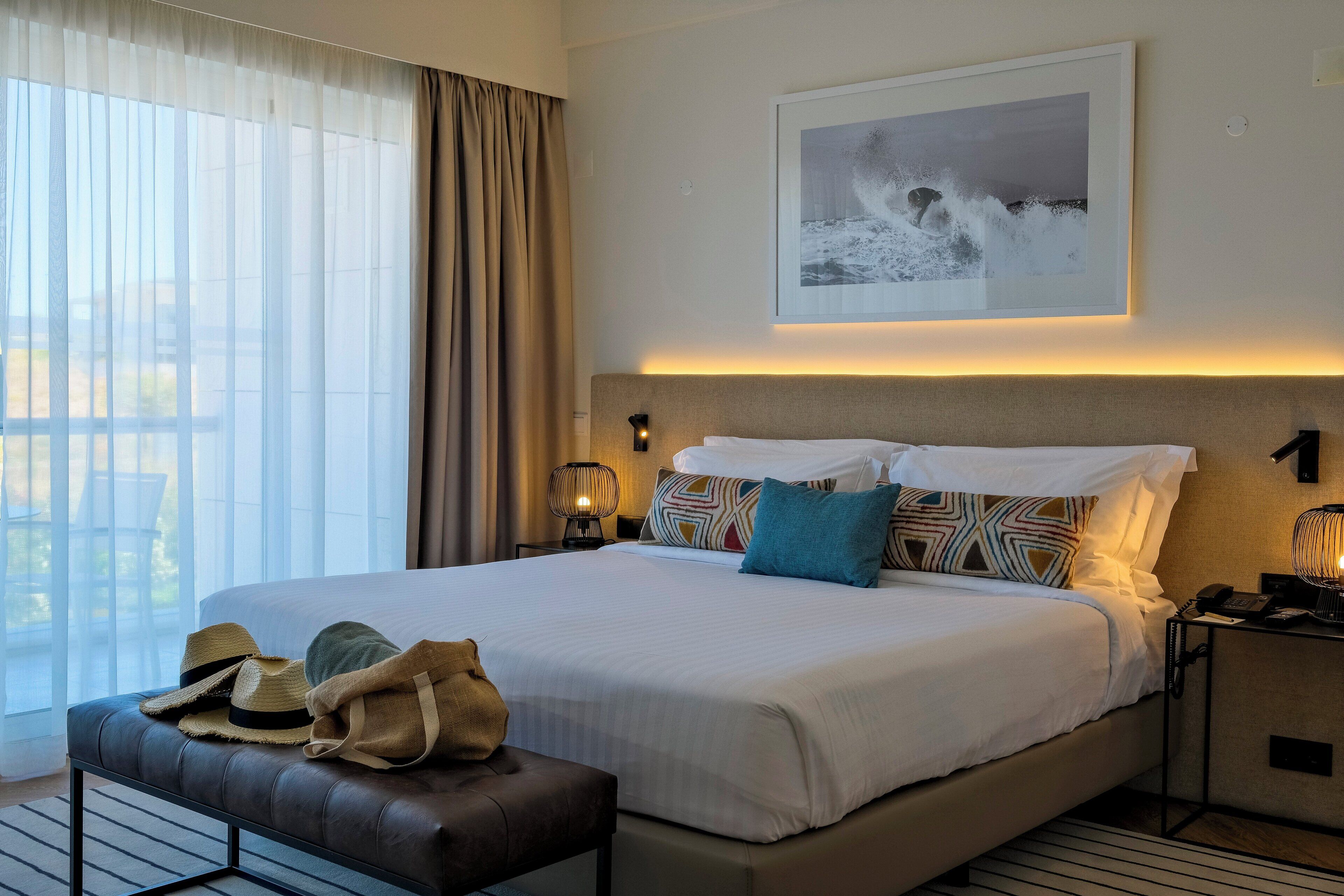A hotel room with a large bed, a bedside table with a lamp, a wall-mounted picture, and a window with curtains.