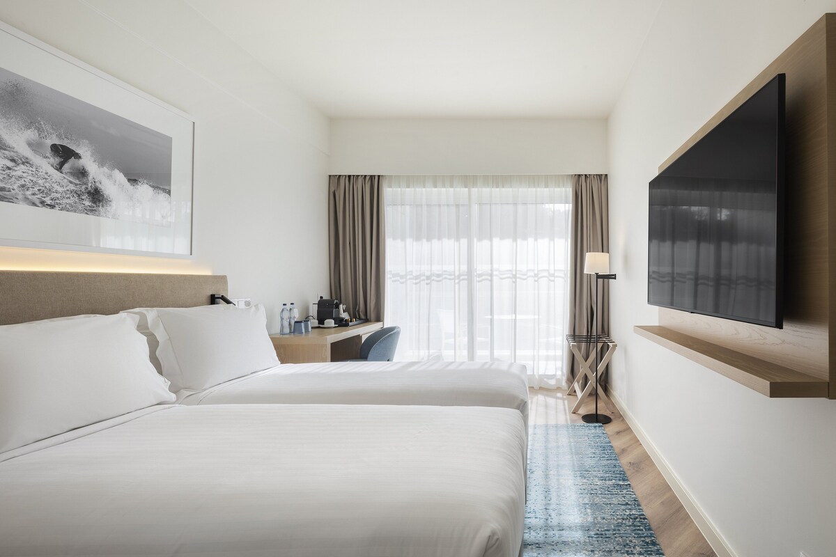 A hotel room with a large bed, a desk with a chair, a flat-screen TV mounted on the wall, and a window with curtains.
