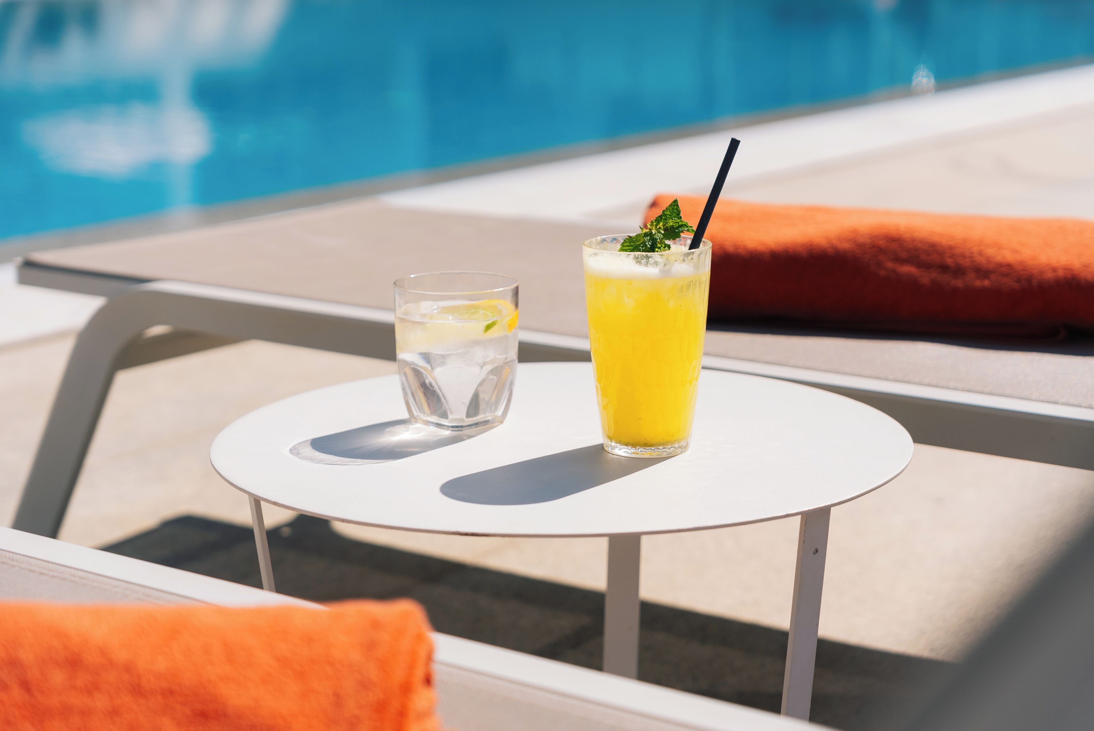 Une table au bord de la piscine avec un verre d’eau et un verre de jus d’orange, une paille et une feuille de menthe en guise de décoration.