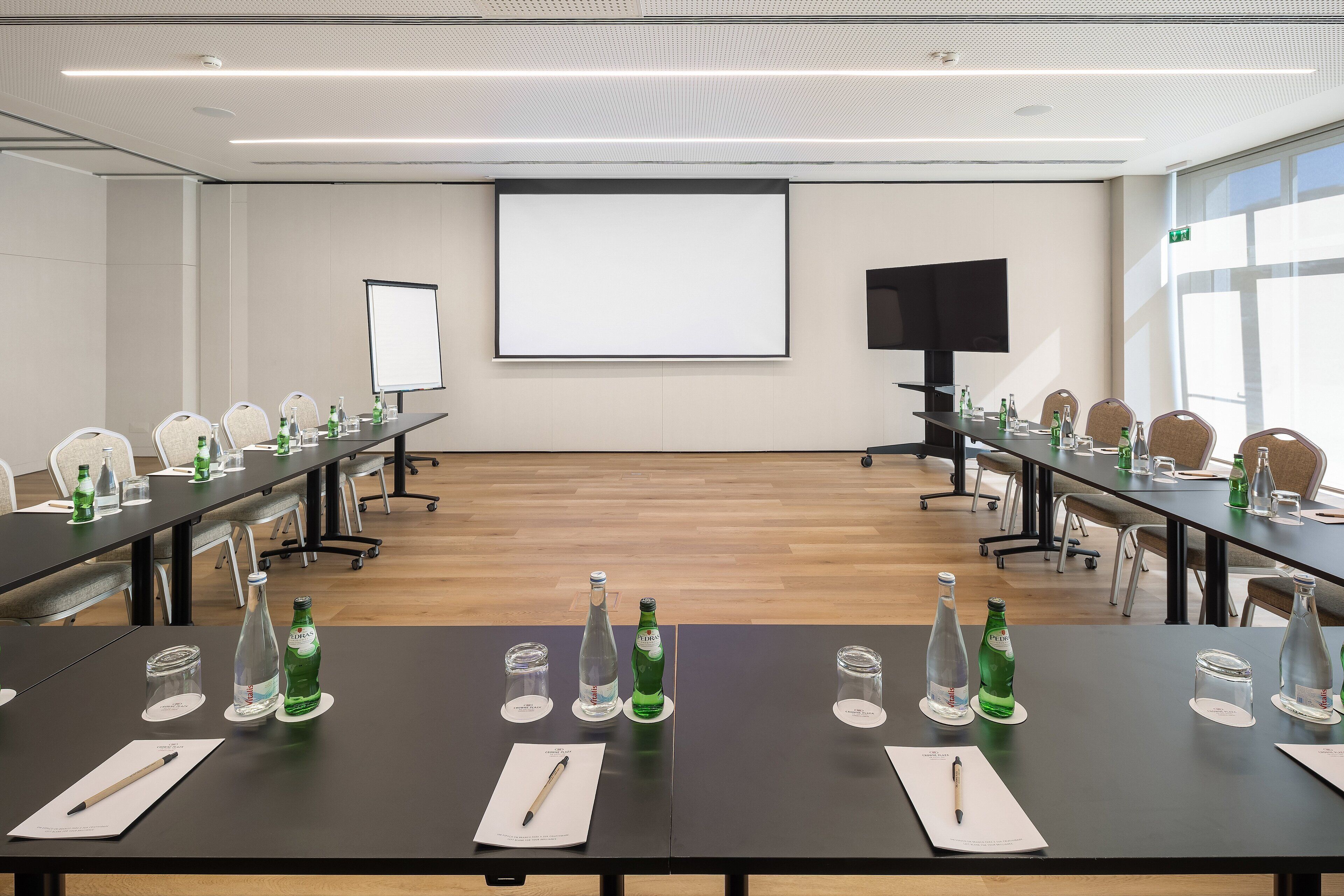 Une salle de conférence équipée de longues tables, de chaises, d’un écran de projection et d’un téléviseur.
