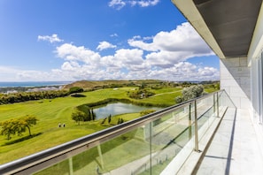 A balcony with a glass railing overlooking a golf course, a pond, and distant hills.