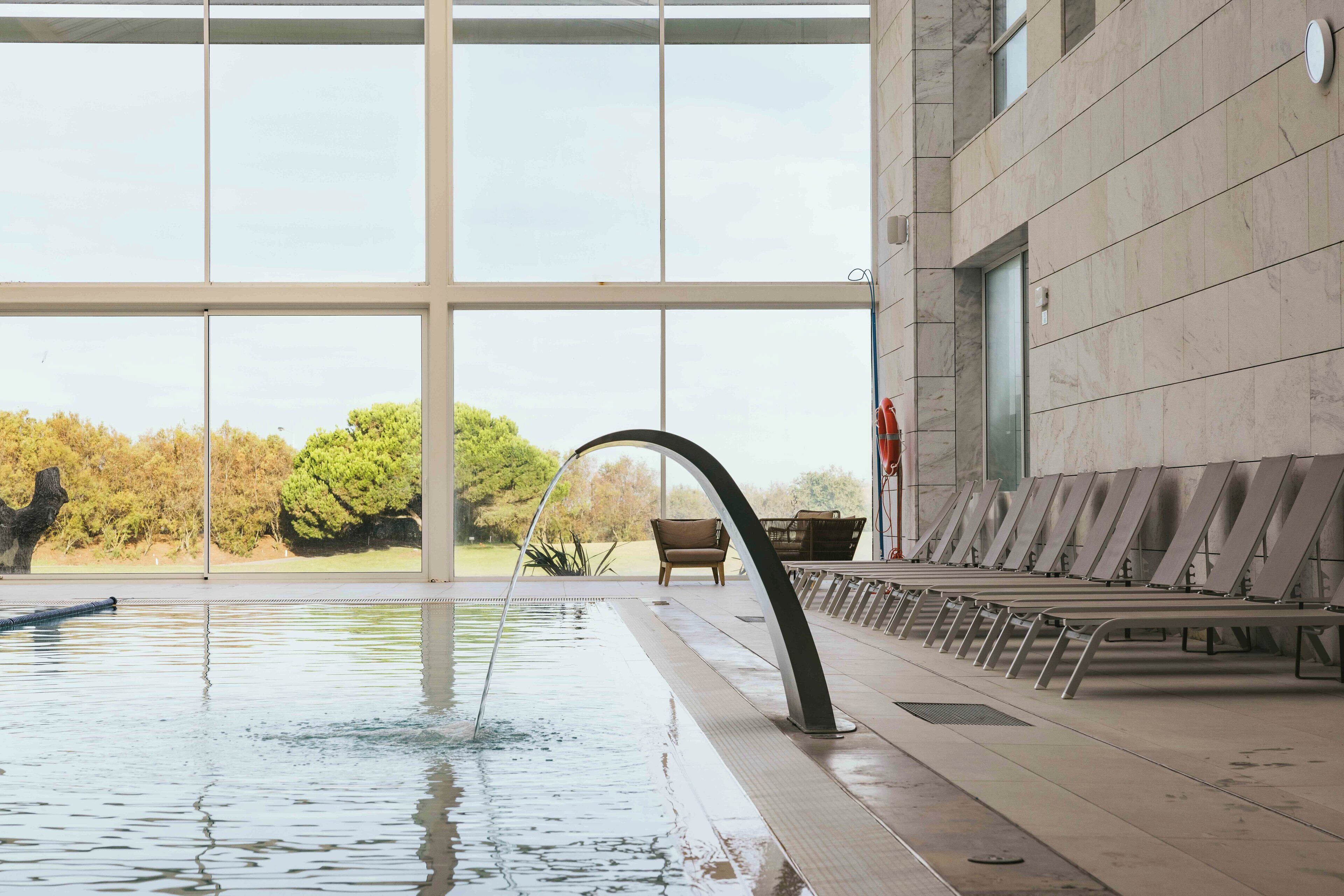 An indoor swimming pool with clear glass walls, a water fountain, and seating along the side.