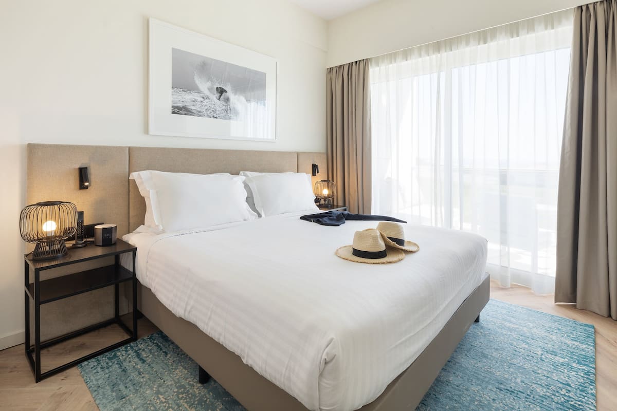 A neatly made bed with white linens, a blue and white striped rug, a framed picture on the wall, and a nightstand with a lamp and a small table.