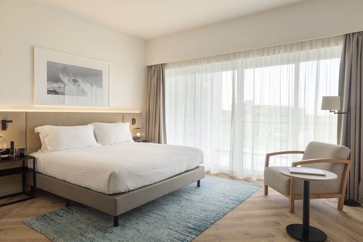 A modern hotel room with a large bed, a side table, a chair, and a large window with sheer curtains.