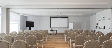 Un salón de conferencias con filas de sillas orientadas hacia una pantalla de proyección, una pizarra y una mesa para presentaciones.