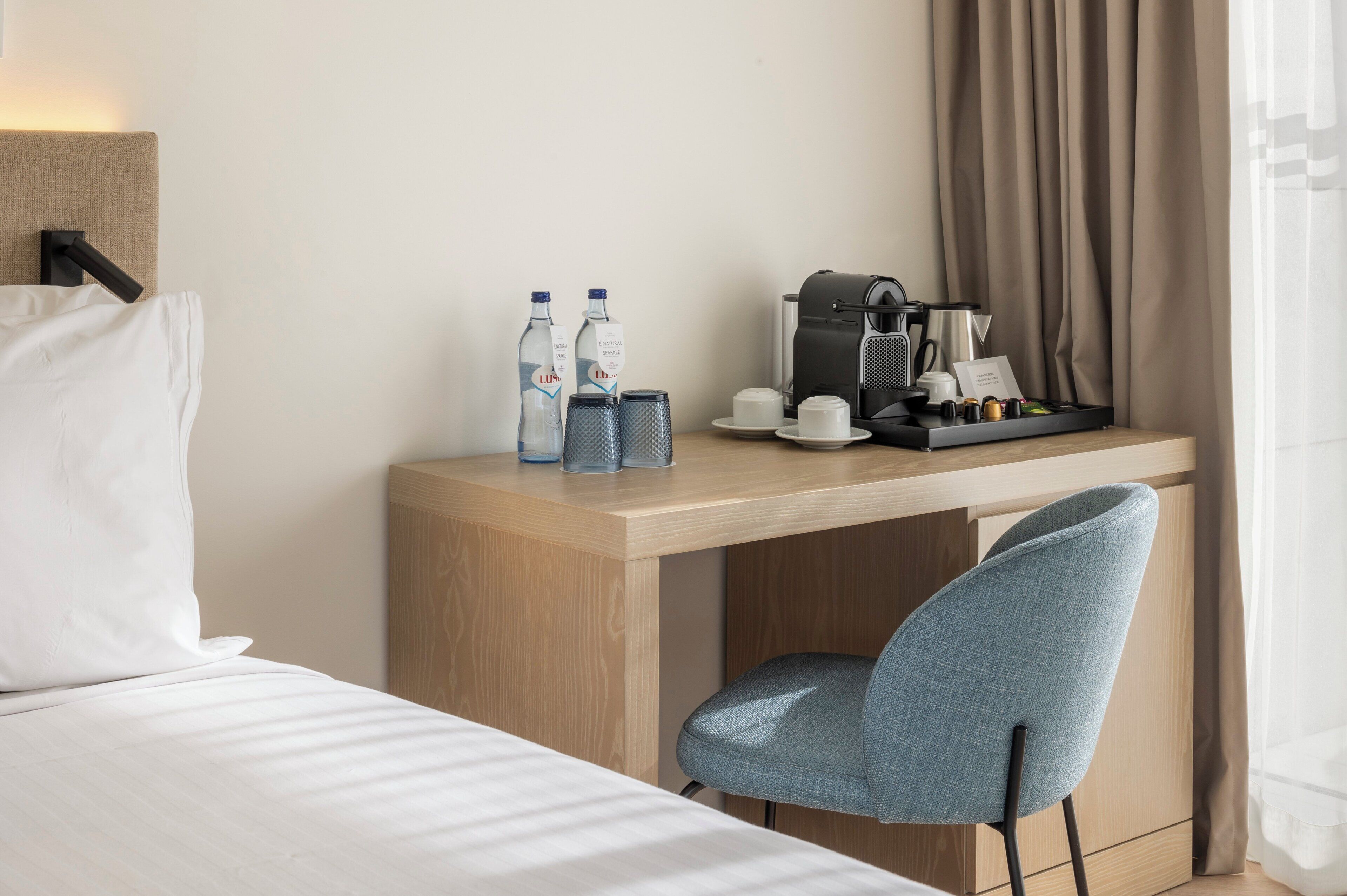 A hotel room with a bed, a desk with a coffee maker, two bottles, and a chair.