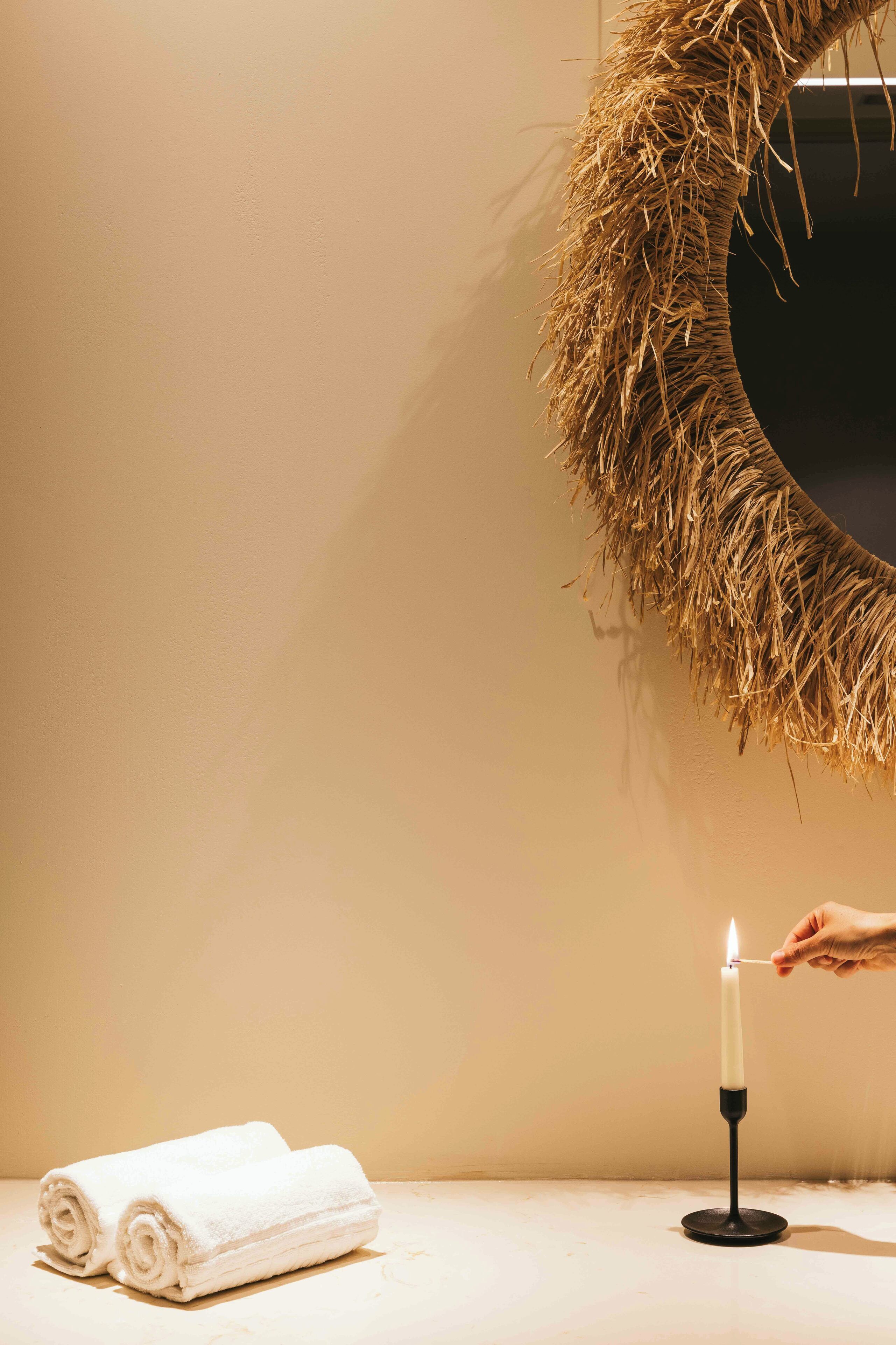 A candle on a black holder, a rolled white towel, and a straw wreath on a wall.