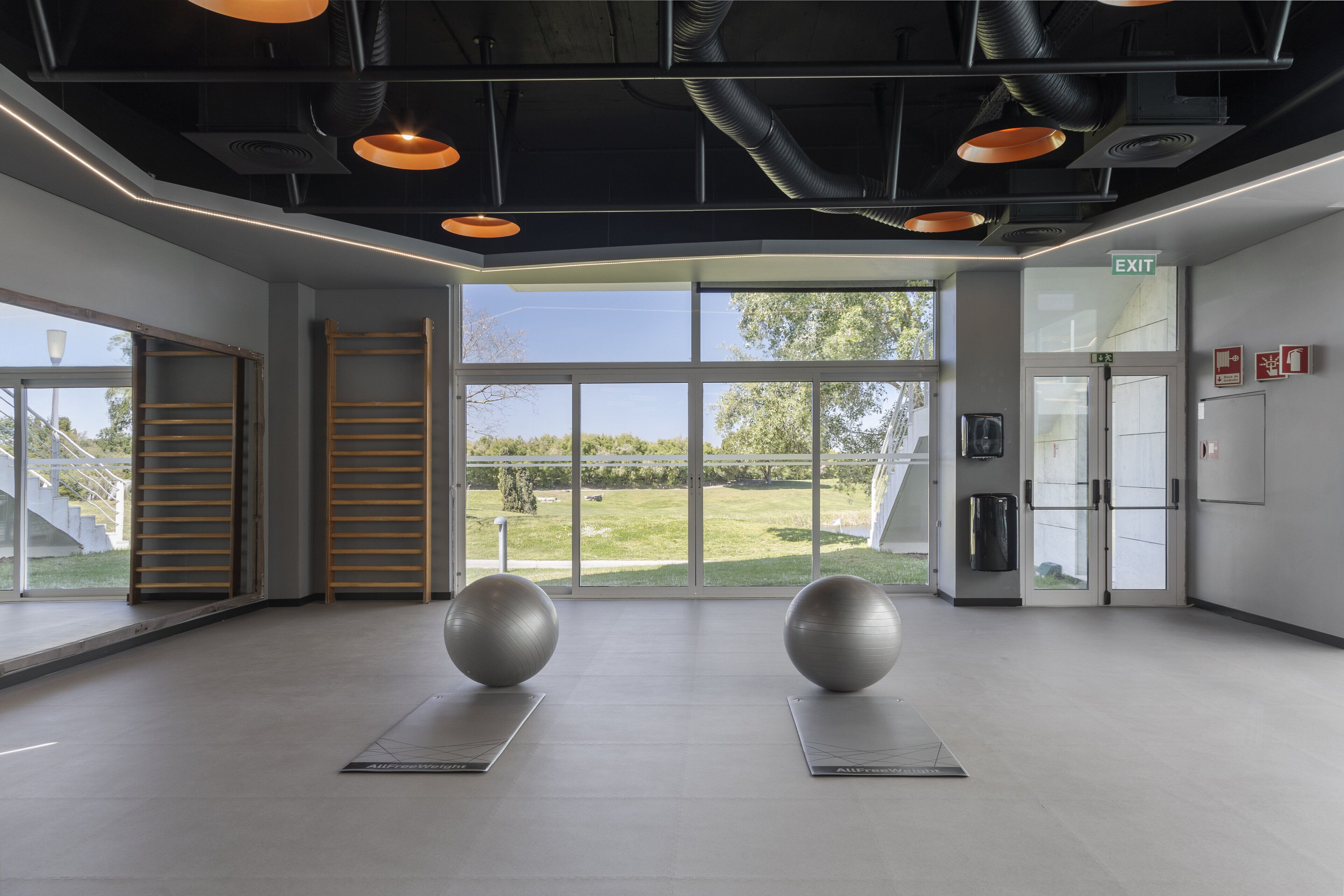 Une pièce spacieuse avec de grandes fenêtres, des ballons d’exercice posés sur des tapis et un panneau indiquant la sortie.