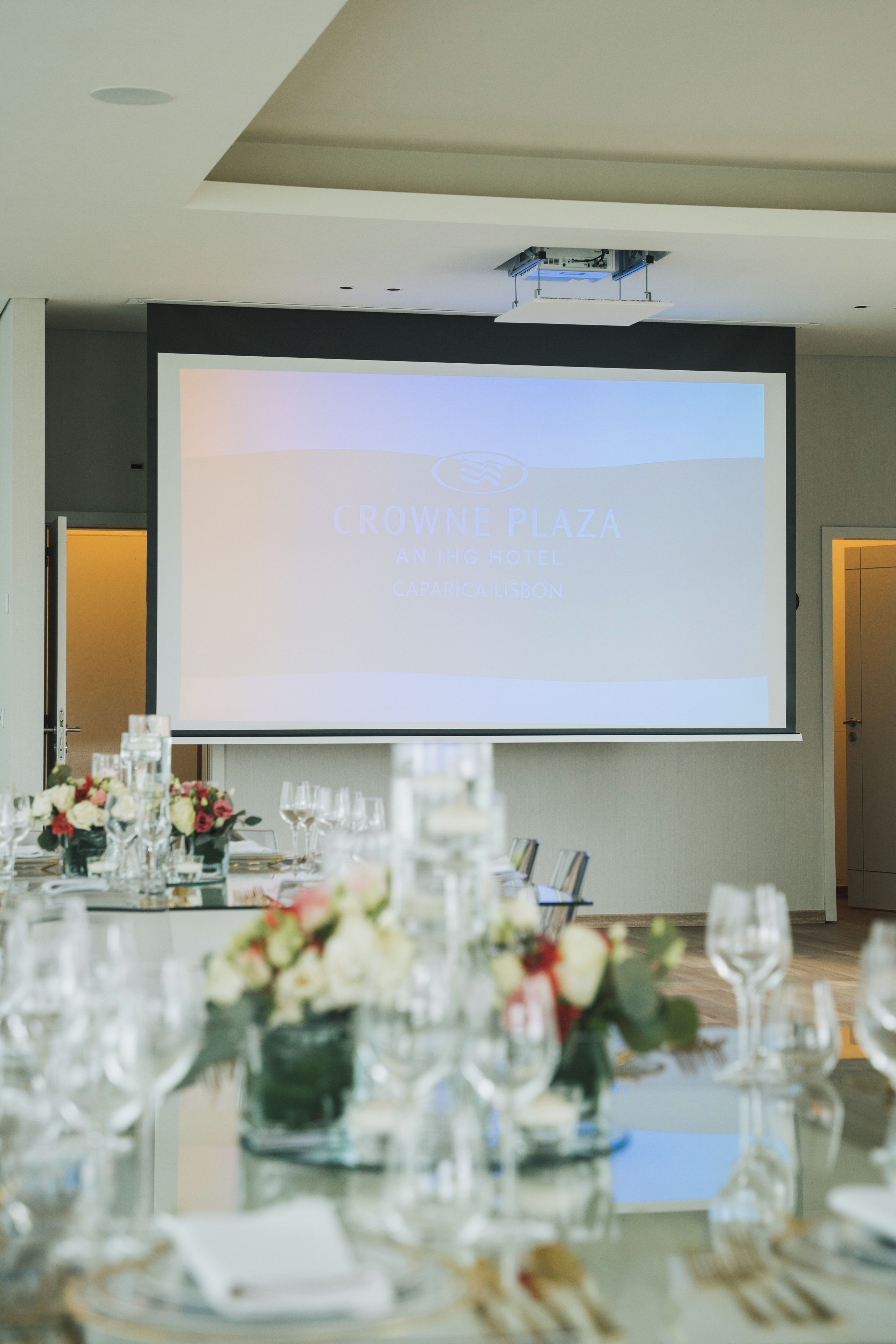 Un salón de conferencias con una pantalla de proyector mostrando 'CROWNE PLAZA AN IHG HOTEL CAPARICA LISBON'.