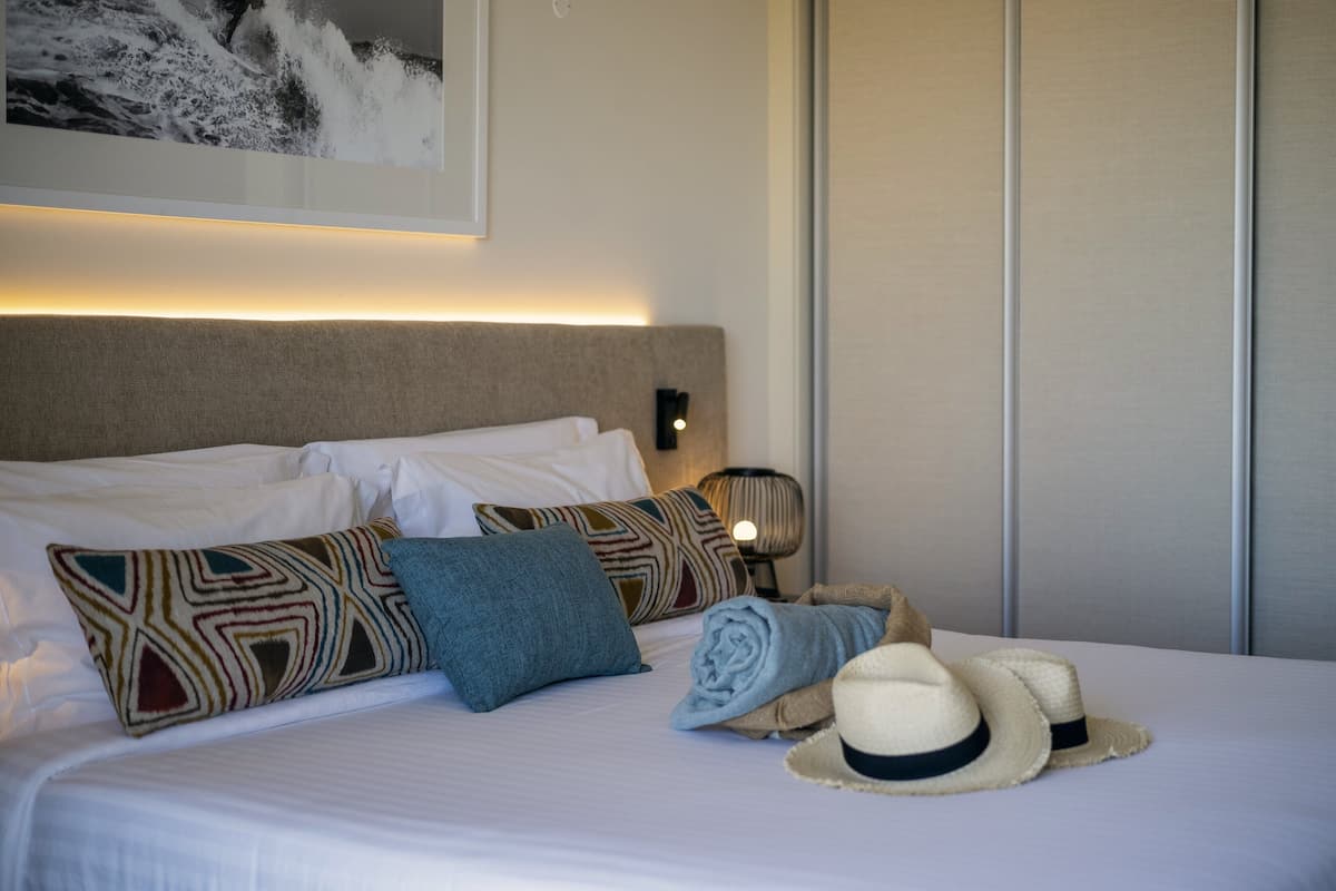 A neatly made bed with patterned pillows, a rolled towel, and hats on the side.