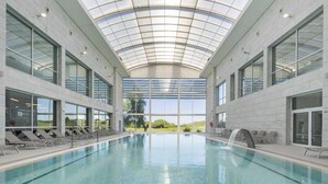 An indoor swimming pool with a glass ceiling, clear views of the outdoors, and sun loungers along the sides. - Crowne Plaza Caparica Lisbon by IHG (Almada)