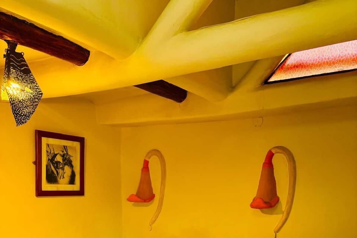 Ein Hotelzimmer mit zwei Betten, jeweils mit weißer Bettdecke und gelben Kissen. An der Wand hängt ein gerahmtes Kunstwerk und über den Betten befindet sich eine außergewöhnliche Leuchte.