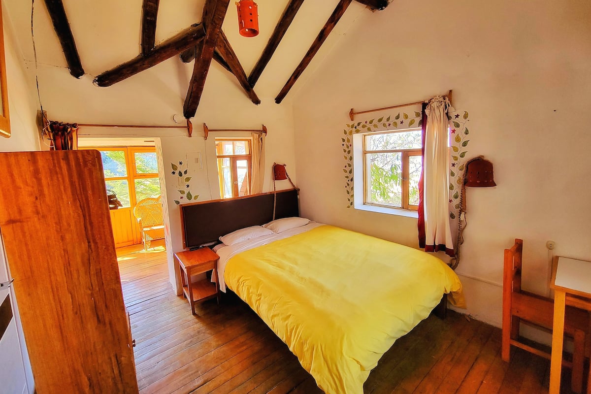 Ein Zimmer mit einem Holzbett, einem Schreibtisch aus Holz, einem Fenster mit Vorhängen und Blick auf Grünfläche.