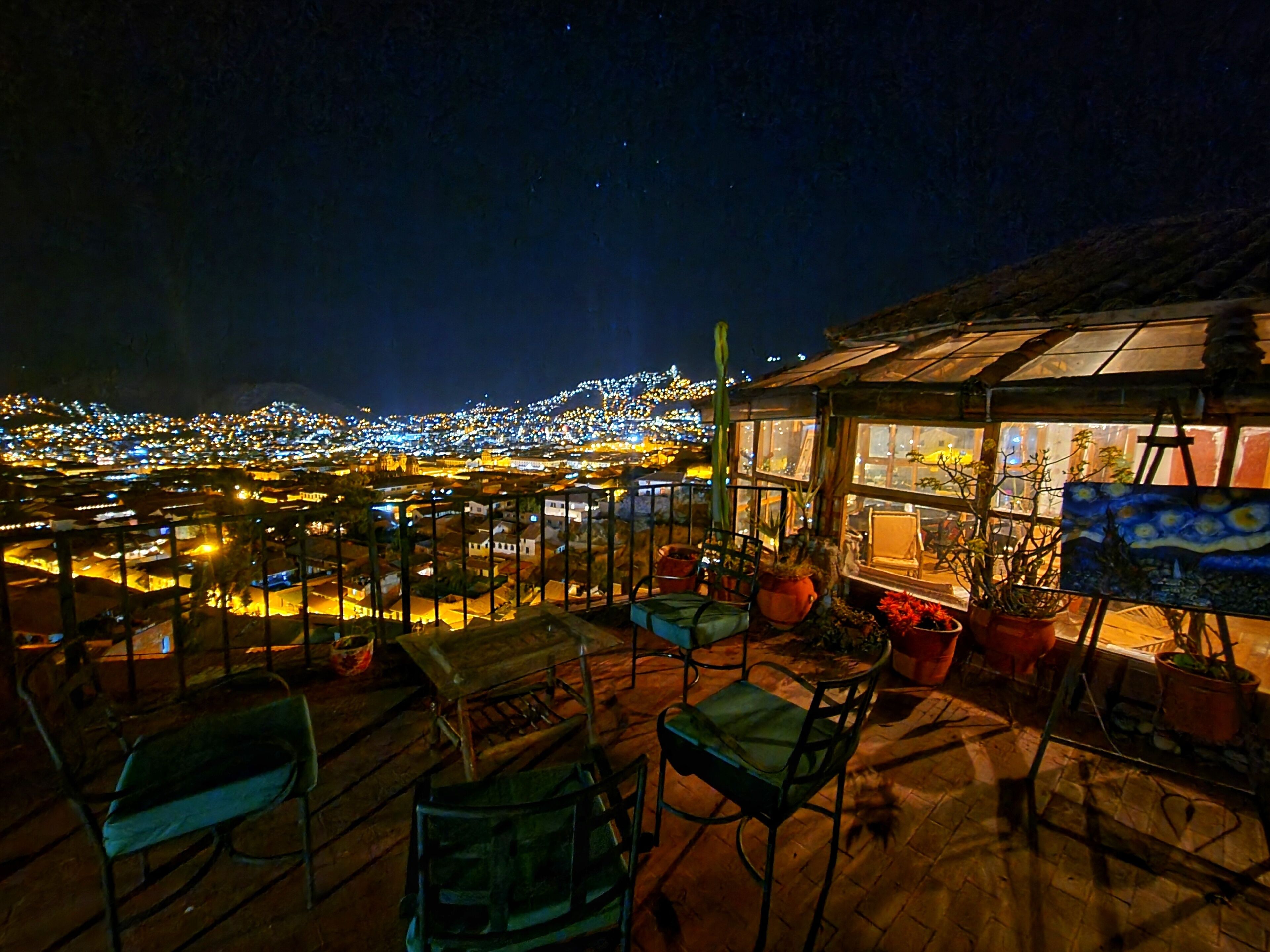 A rooftop terrace with chairs and potted plants, overlooking a cityscape at night.