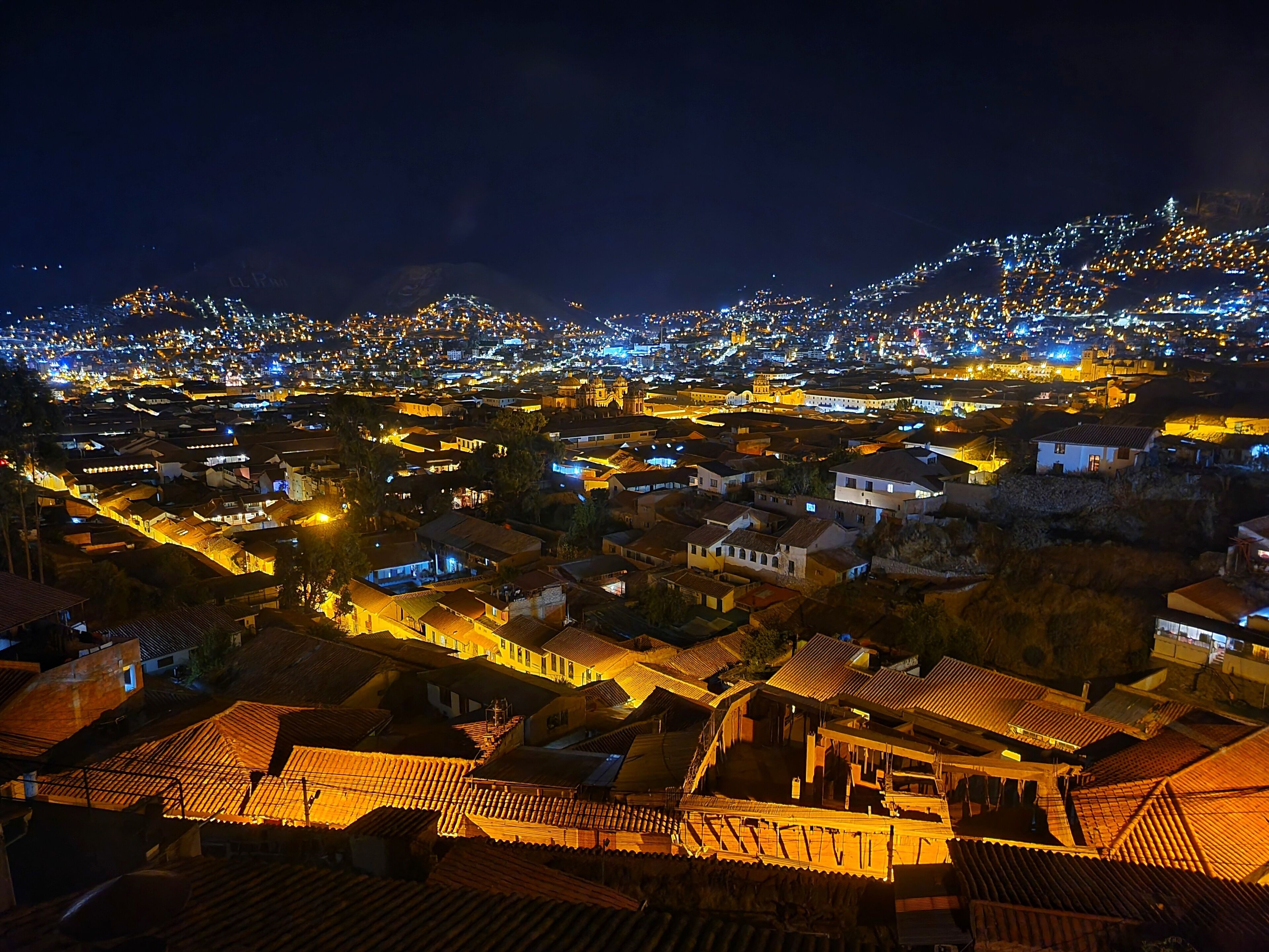 A nighttime view of a town with illuminated rooftops and buildings, indicating a densely populated area with historical architecture.
