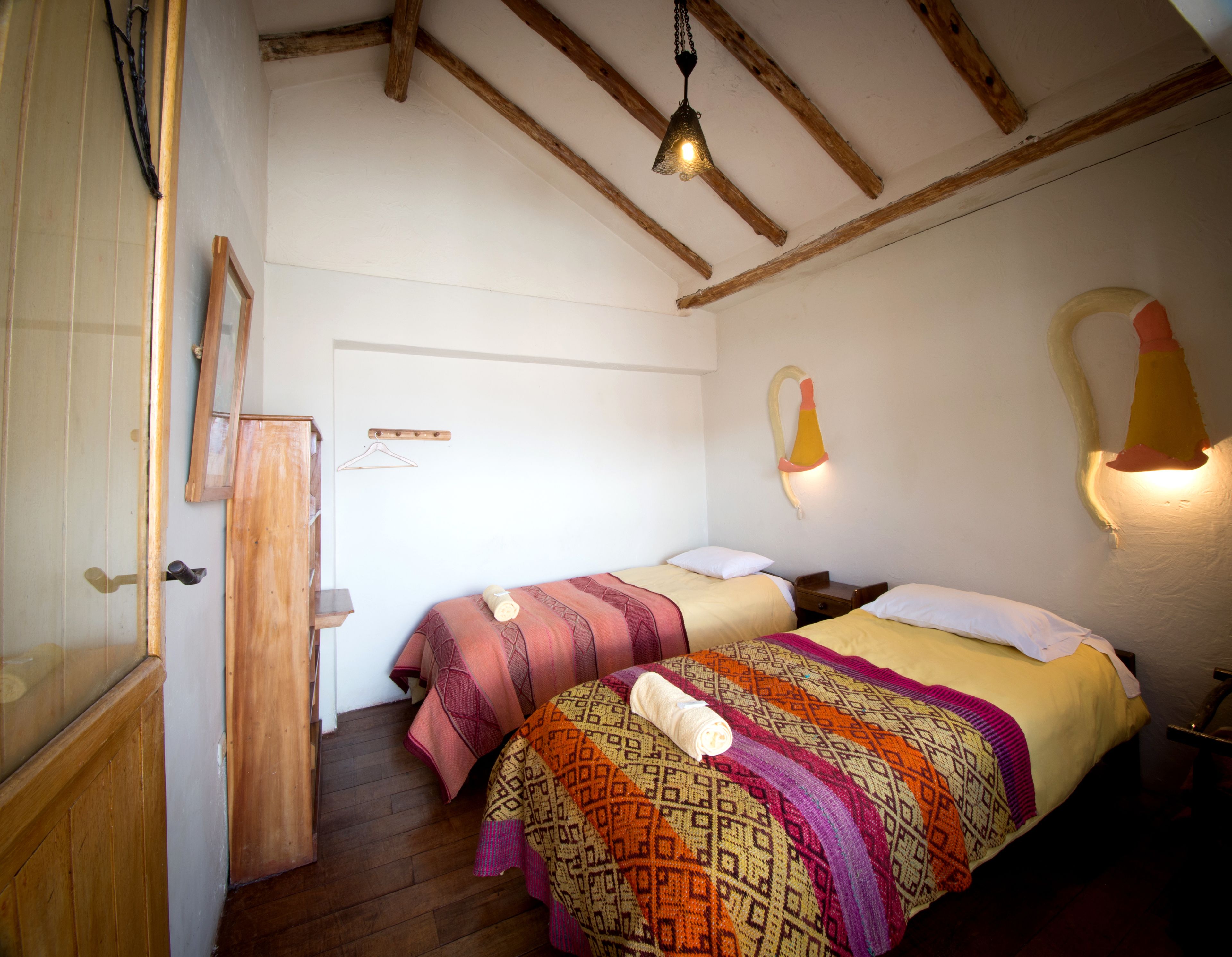 A room with two beds, wooden beams on the ceiling, a hanging light fixture, and a wooden door.