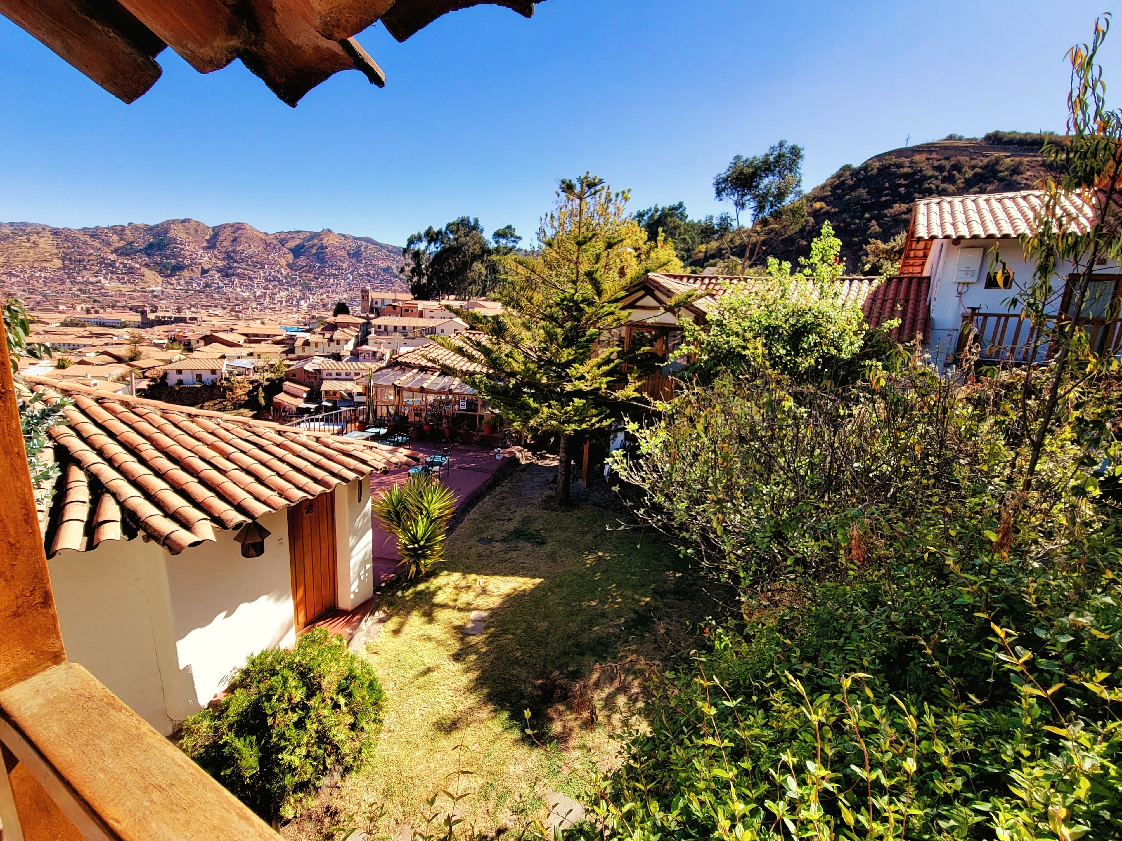 A residential area with terracotta-roofed houses, a small garden, and a view of a distant town and hills.
