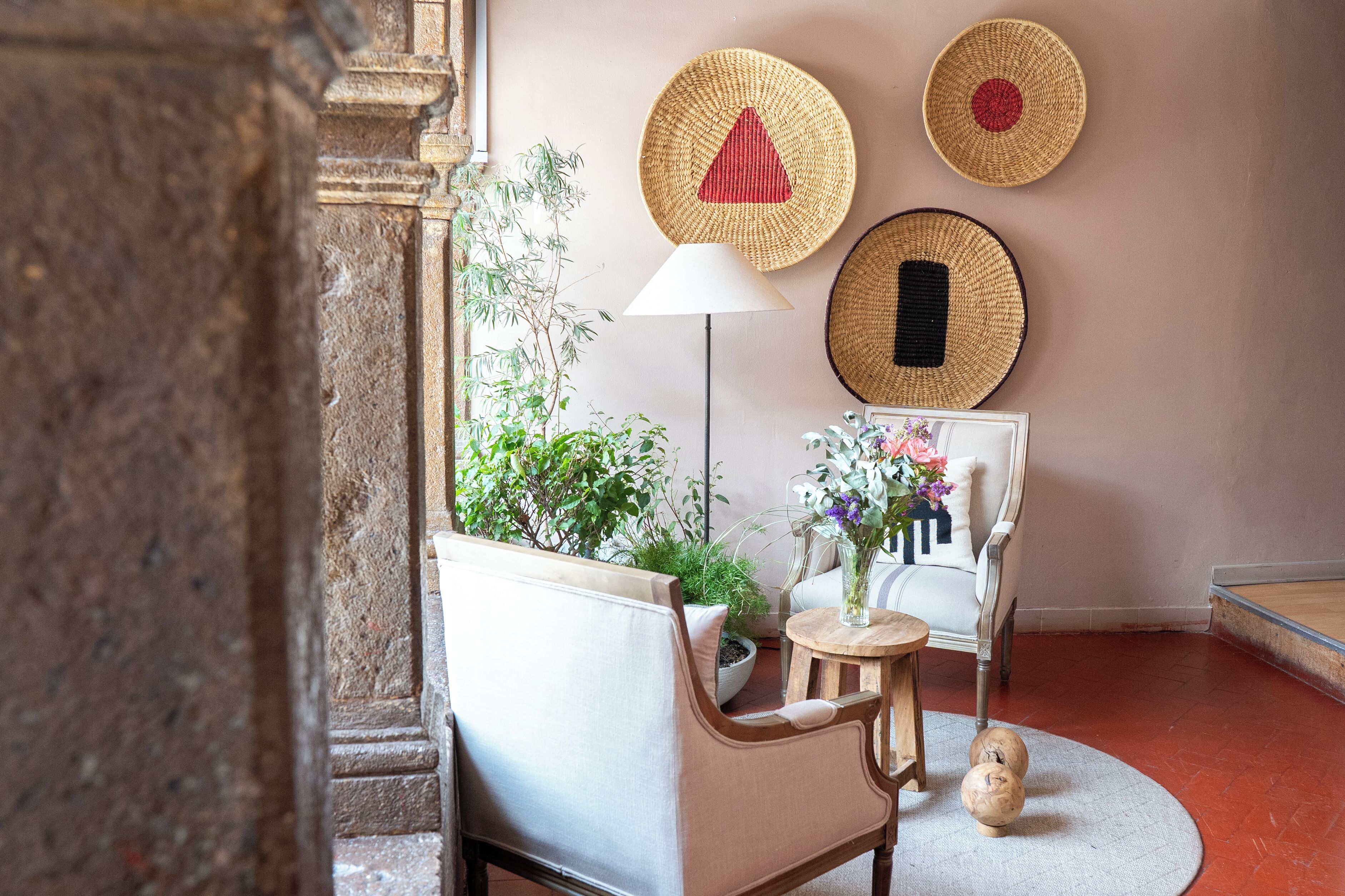 Un cuarto acogedor con un sillón blanco, una pequeña mesa redonda con un florero y una lámpara de pie. Hay tres cestas tejidas en la pared y una planta en maceta junto al sillón.