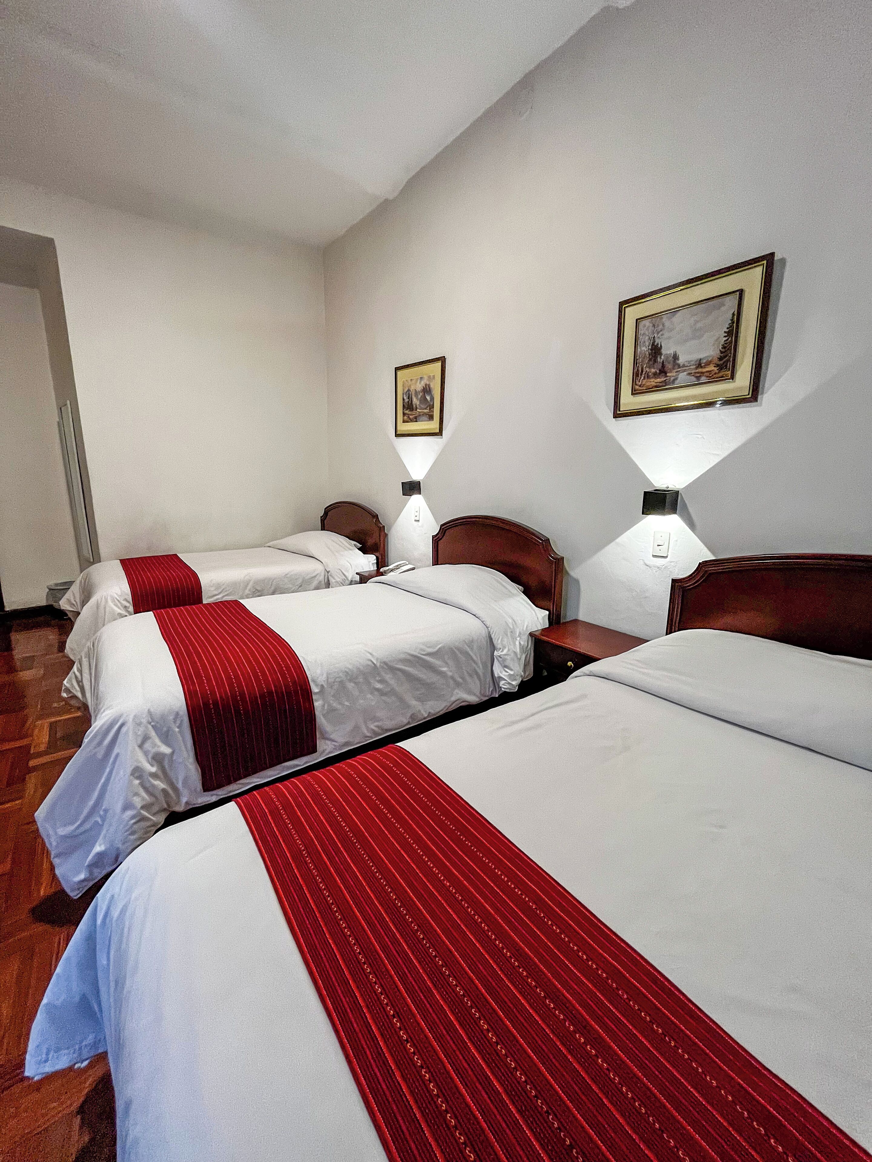 Habitación de hotel con tres camas, cada una con ropa blanca y detalles rojos. Hay dos lámparas de pared y un cuadro enmarcado sobre las camas.