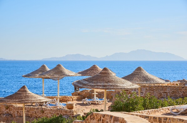A coastal resort with stone walls, thatched umbrellas, and lounge chairs by the sea.