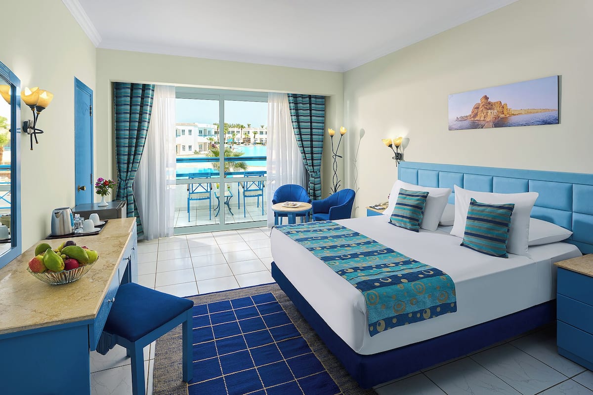 Quarto de hotel com cama, escrivaninha com fruteira, cadeira e vista para piscina e prédios lá fora.