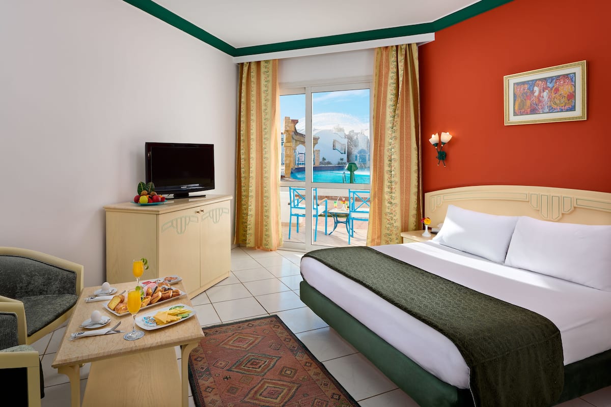 Quarto de hotel com cama, televisão, mesa de jantar com café da manhã, vista para a piscina e um quadro na parede.