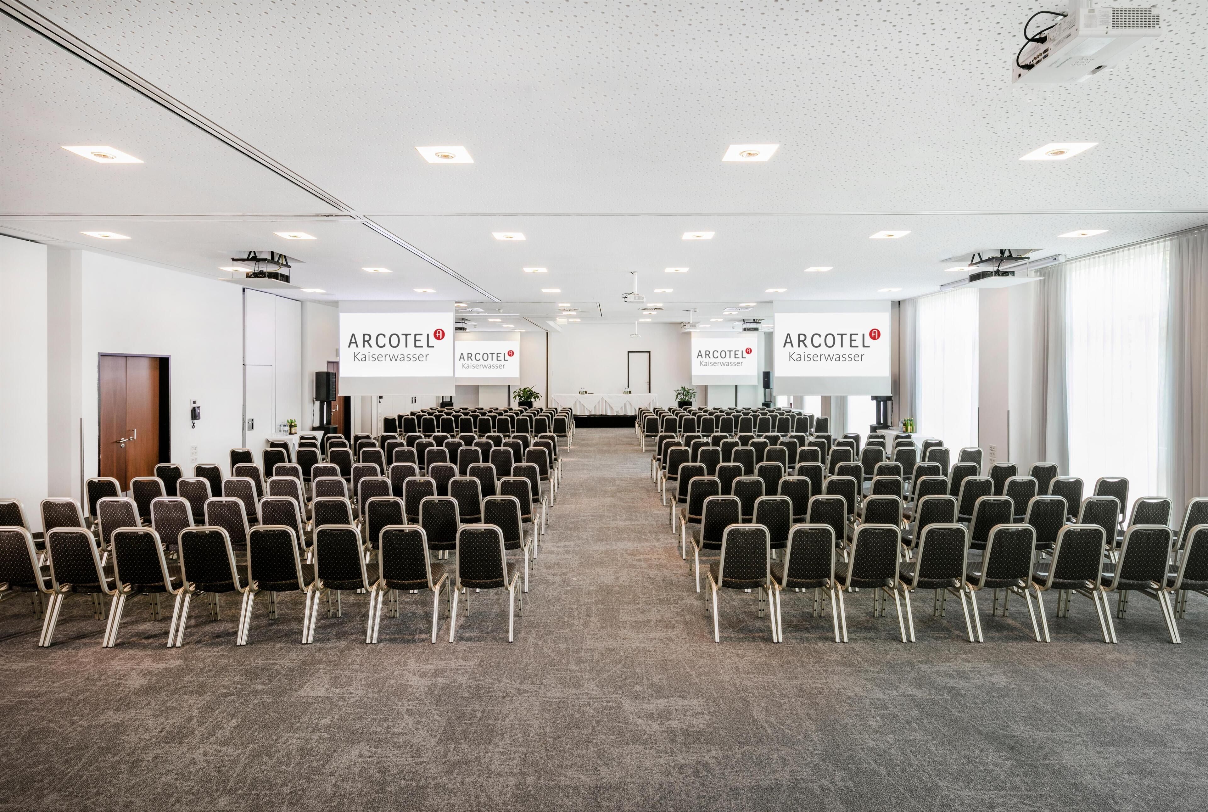 Een vergaderzaal met rijen stoelen, witte muren en plafondprojectoren.