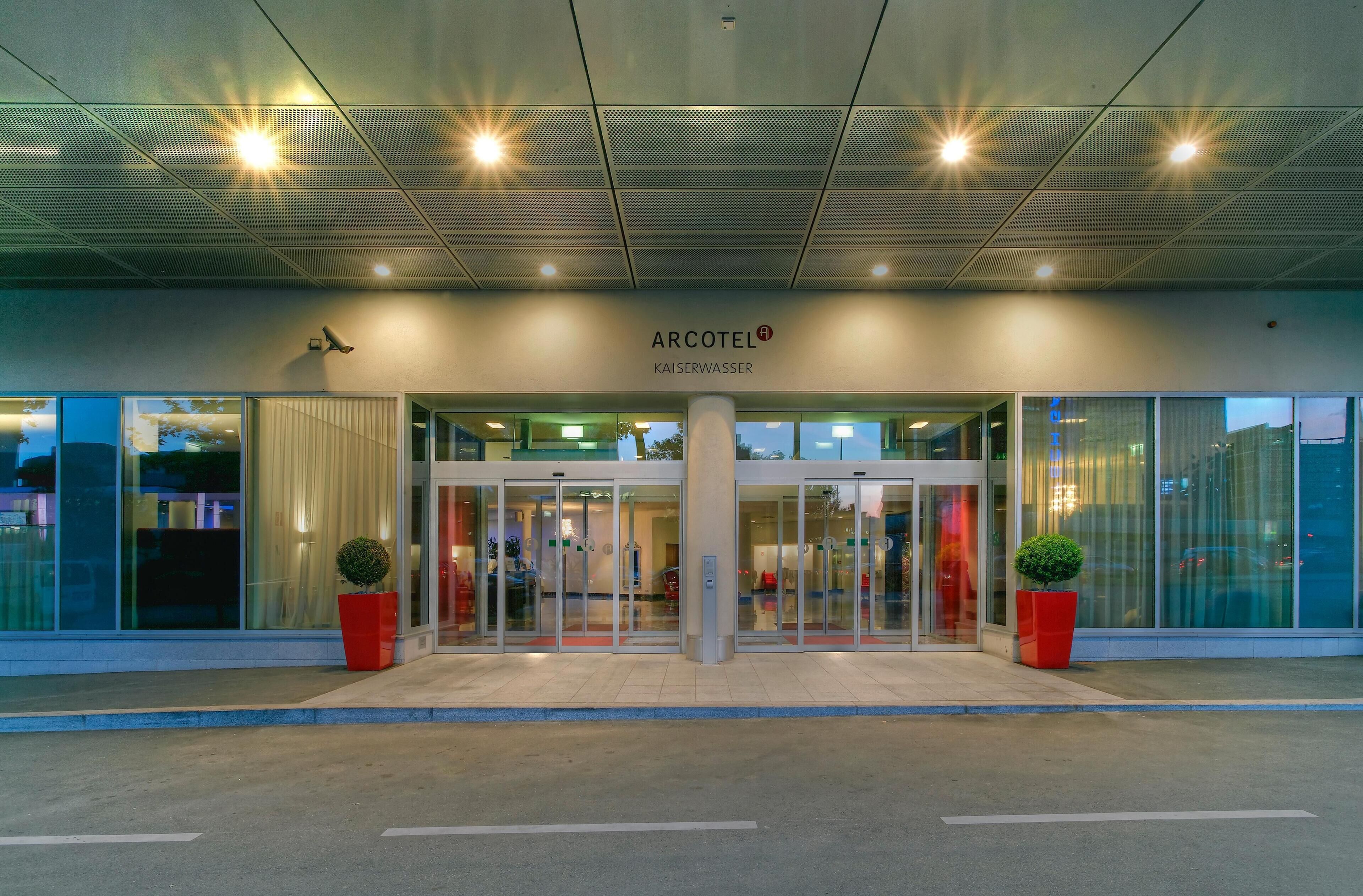 A hotel entrance with a modern design, featuring a glass facade, a central pillar, and red planters.