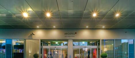 A hotel entrance with a modern design, featuring a glass facade, a central pillar, and red planters.