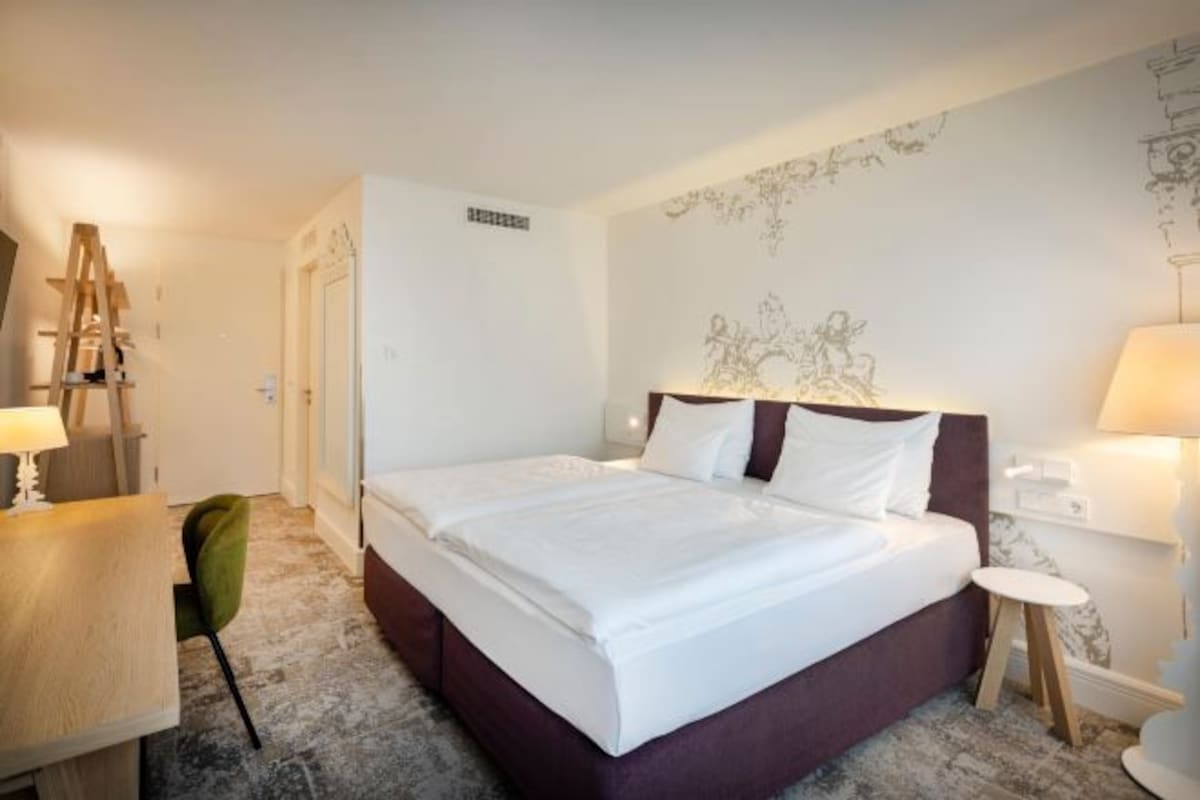 A hotel room with a bed, bedside table, lamp, and a desk with a chair.