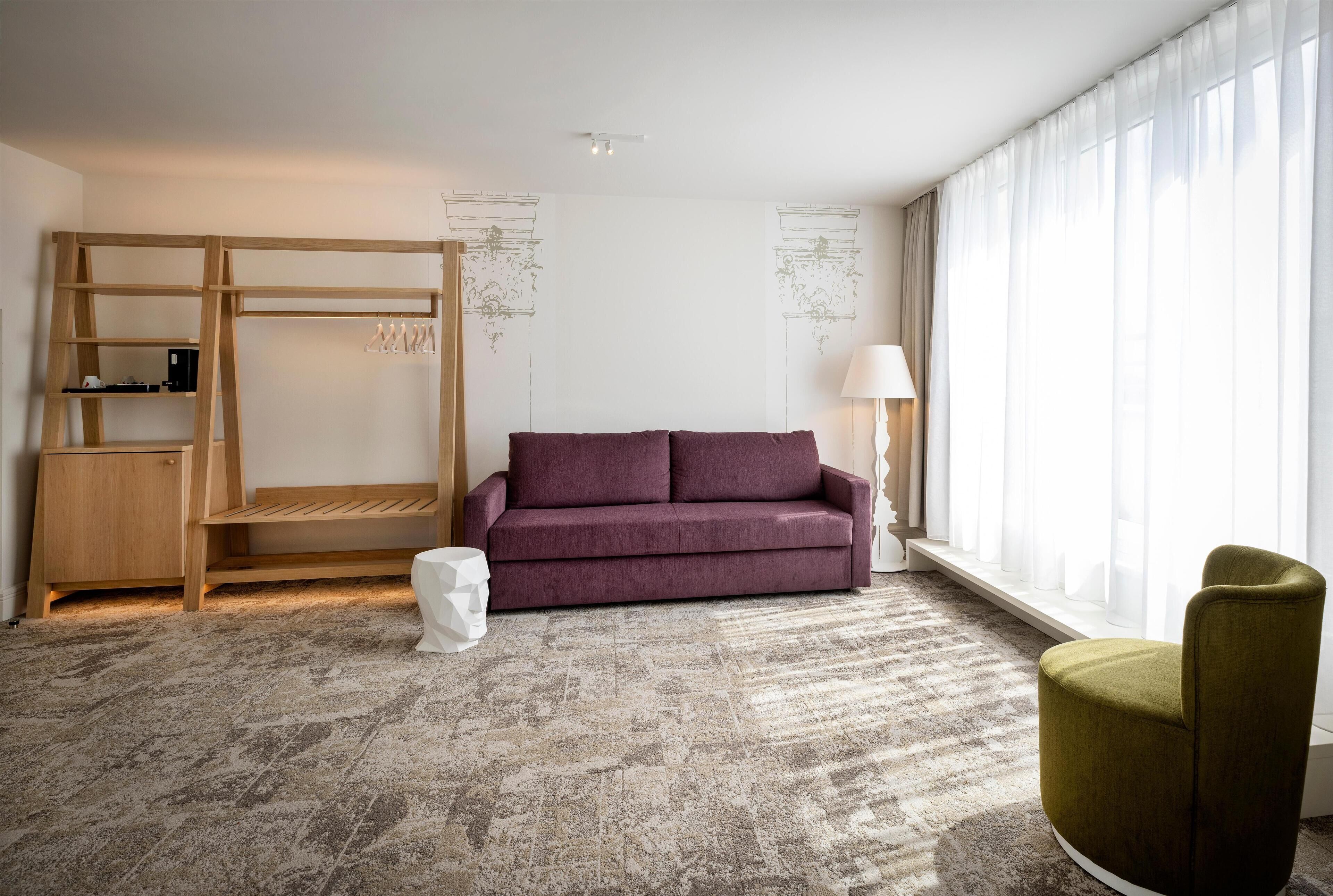 A modern room with a wooden bunk bed, a purple sofa, a green armchair, and a large window with sheer curtains.